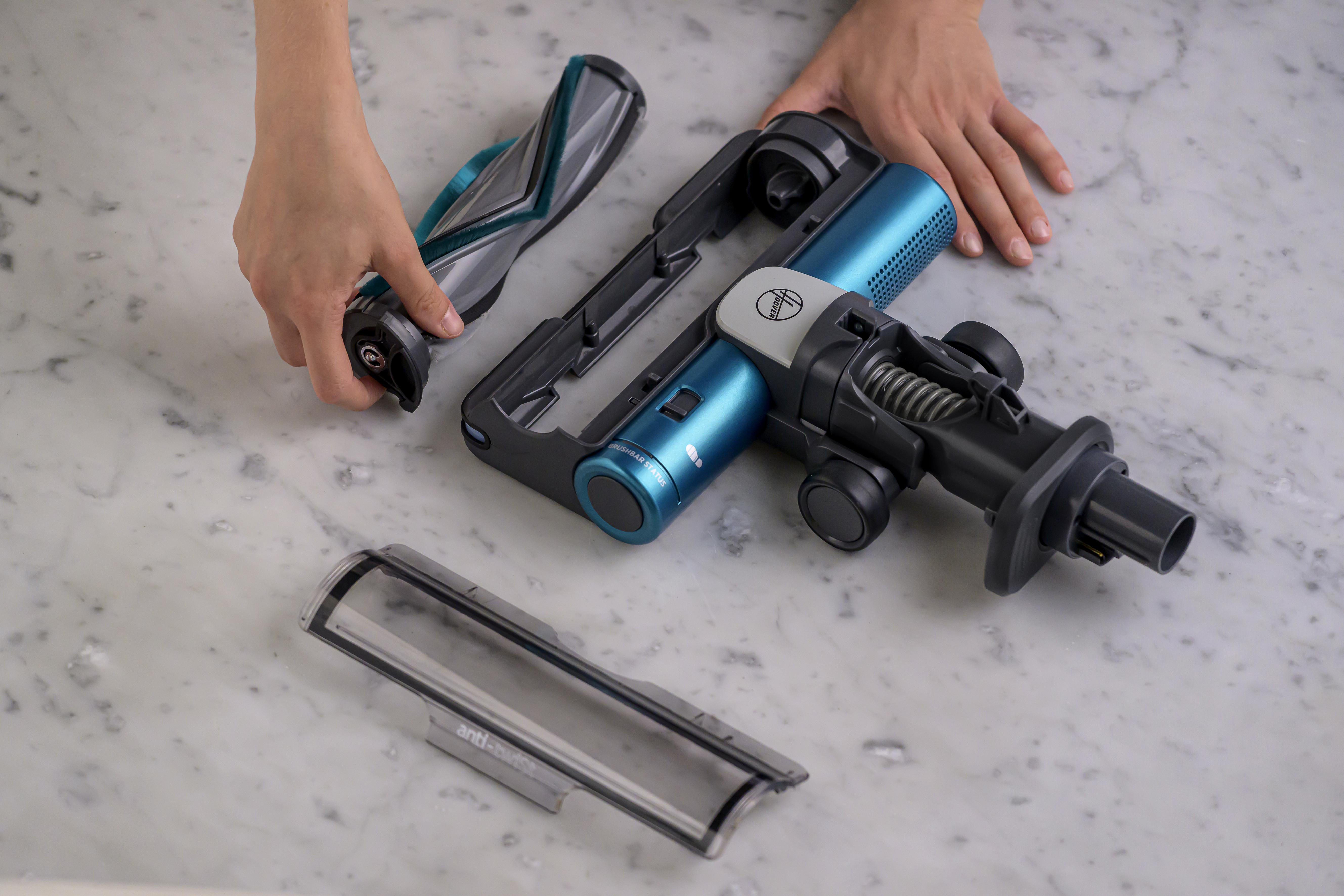 Hände zerlegen einen Staubsauger. Die Teile umfassen eine Walzenbürste, einen blauen Motor und eine transparente Abdeckung.