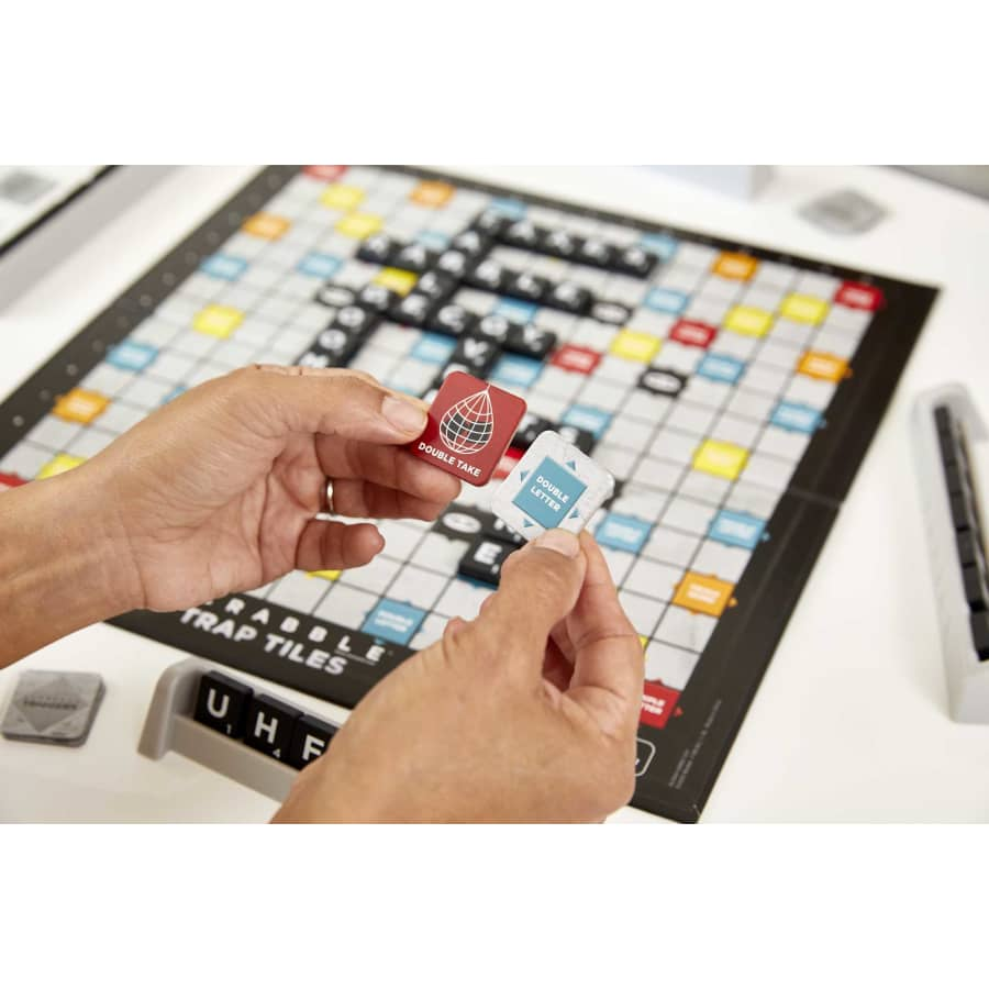 Hände halten zwei Spielsteine über einem Scrabble-Brett. Einer sagt 'Double Take', der andere 'Double Letter'.