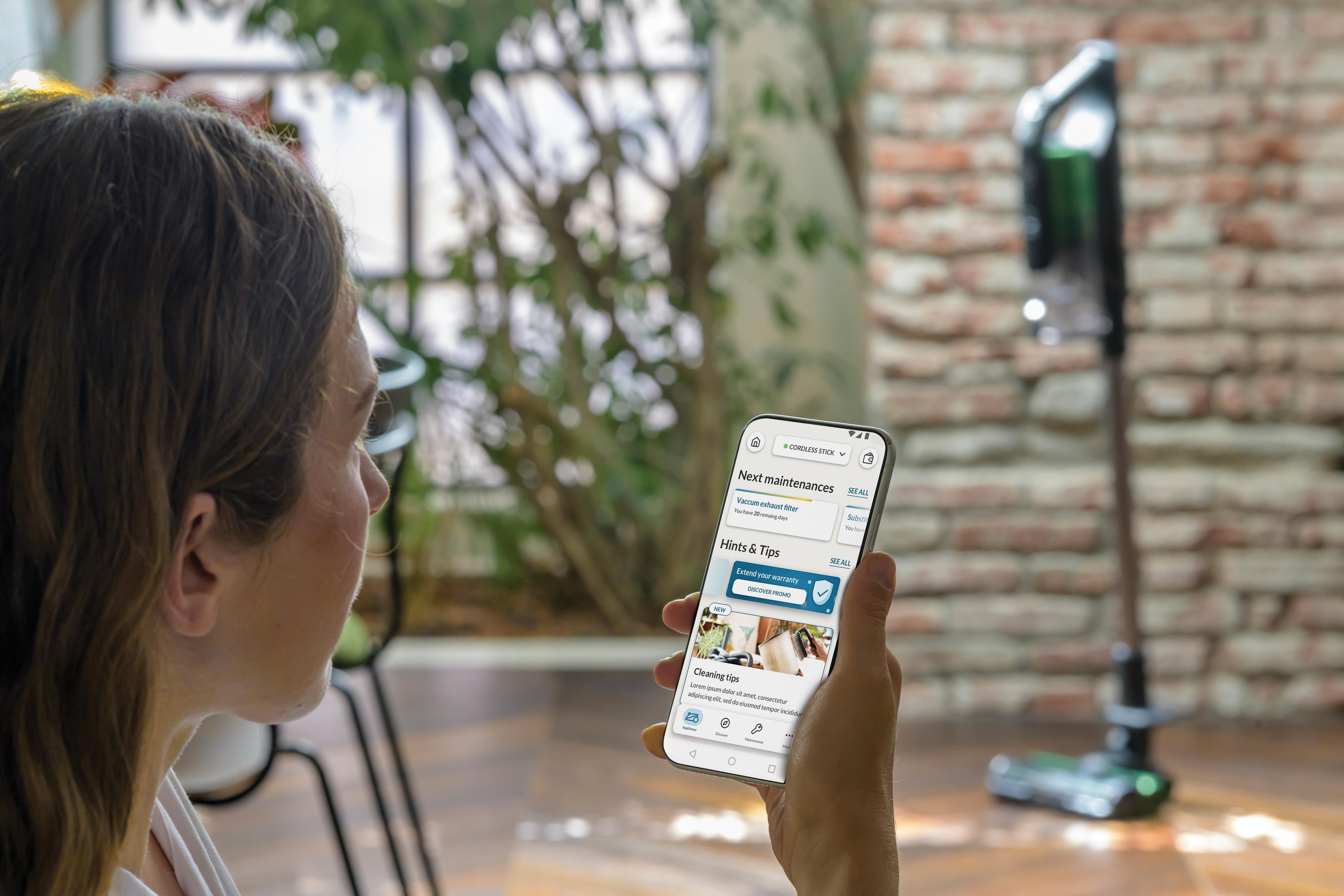 Frau, die auf einen Telefonbildschirm mit einer App schaut. Ein Staubsauger ist im Hintergrund unscharf.