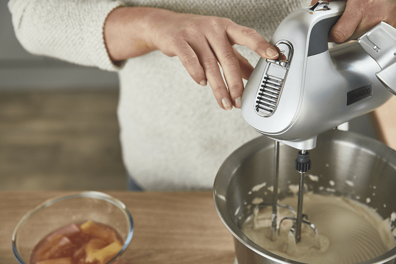 Egy személy a Kenwood ezüst kézi mixerét használja. Egy tálnyi hozzávaló van a közelben.