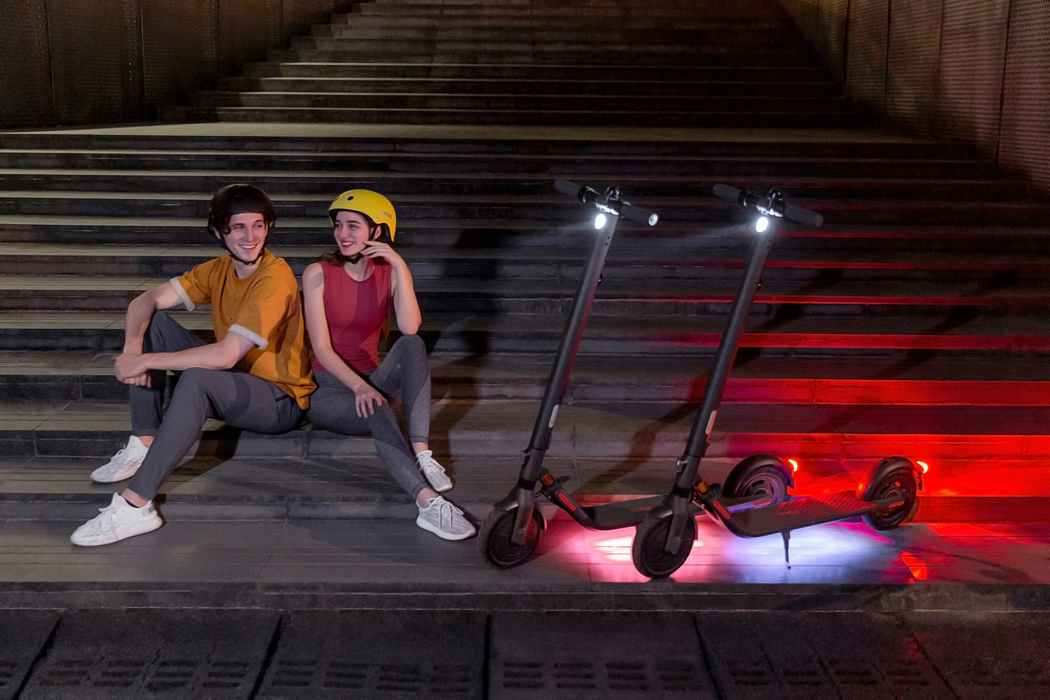 İki kişi merdivenlerde scooterlarla oturuyor. Işıklarla gece sahnesi.