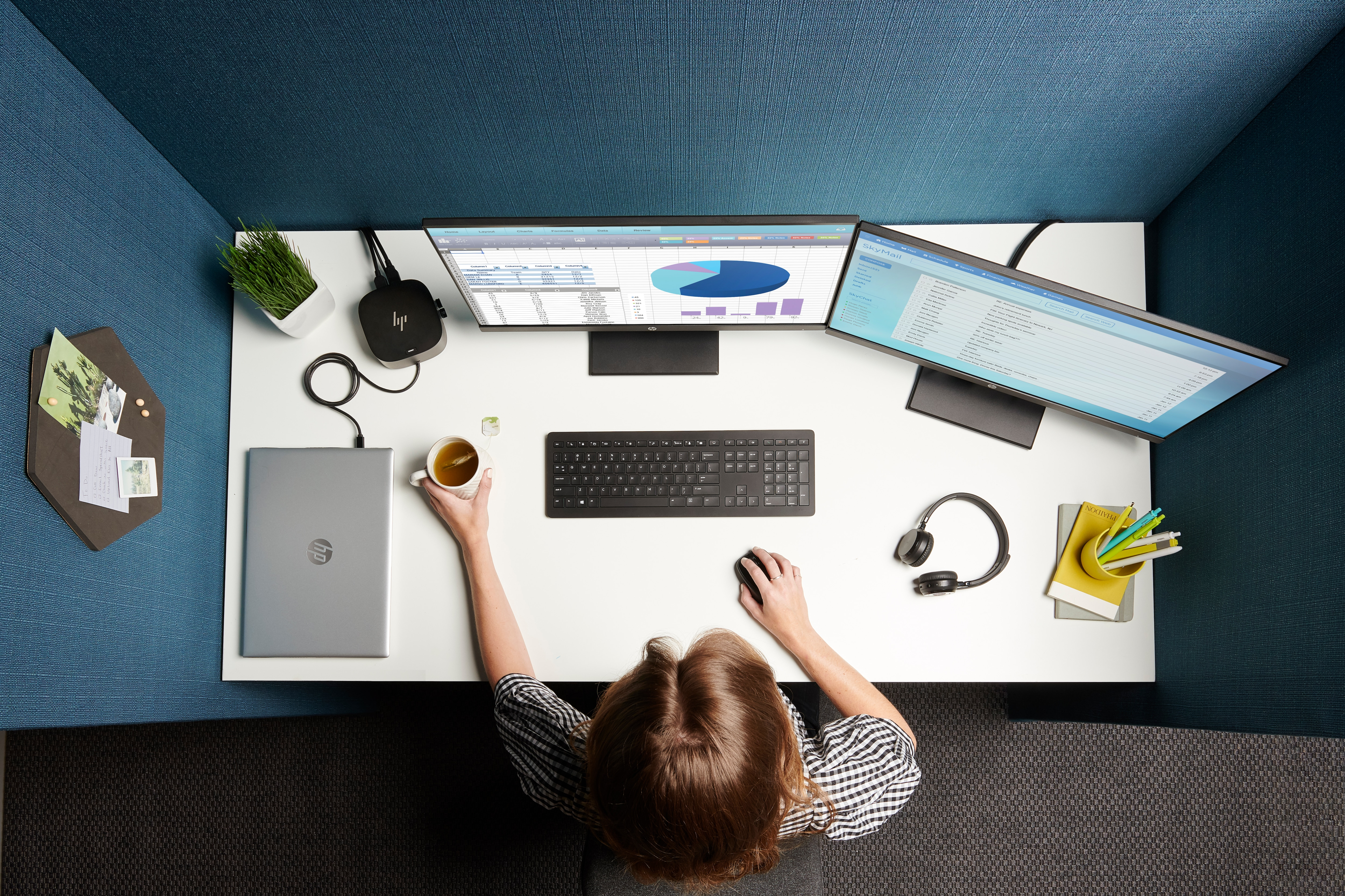 Draufsicht auf einen Schreibtisch mit zwei Monitoren, einem Laptop und einer Person. Zubehör und Pflanzen vorhanden.