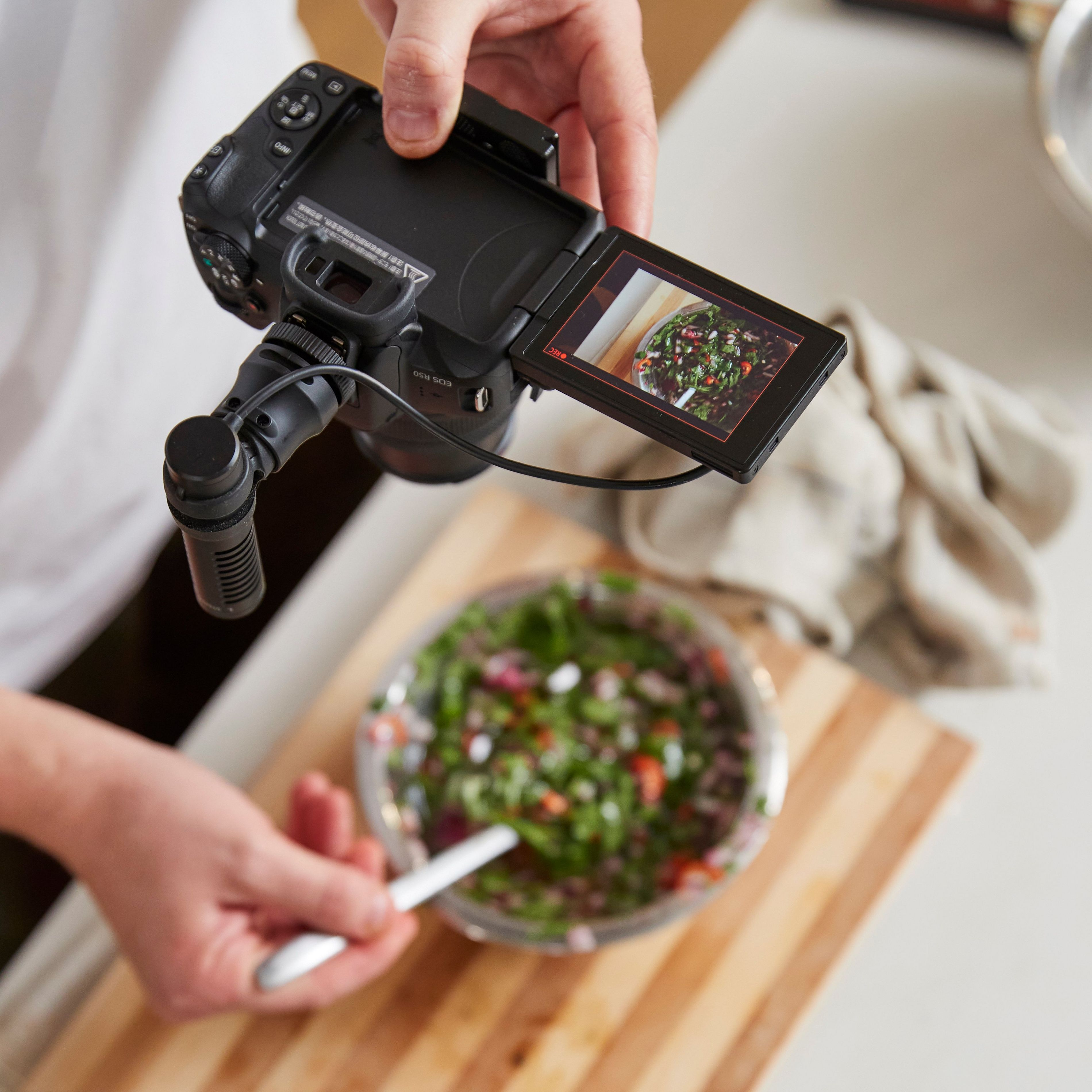 Eine Person filmt einen Salat mit einer Kamera. Die Kamera zeigt den Salat auf einem Holzbrett.