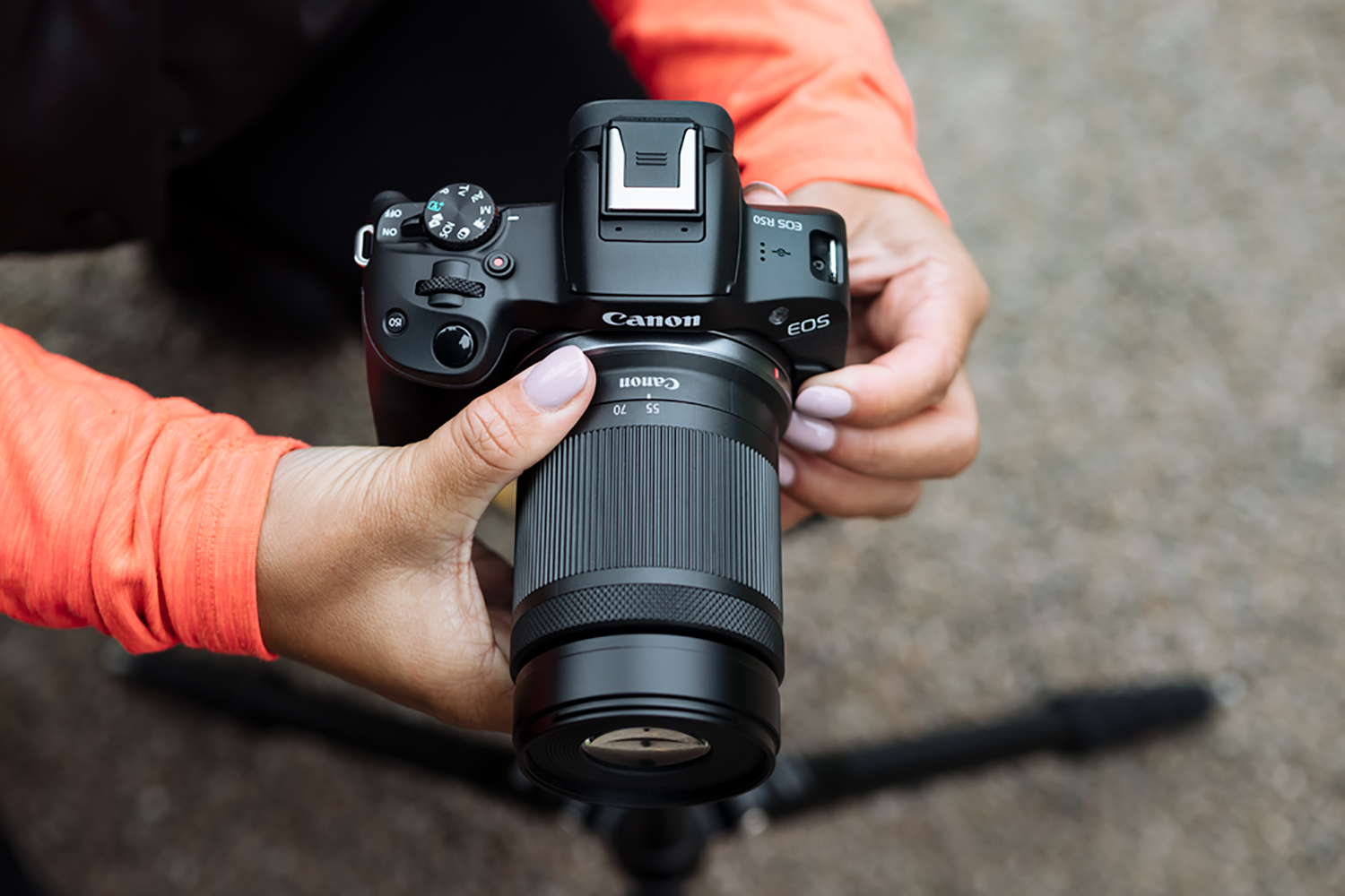 Handen die een zwarte Canon EOS R5 camera vasthouden. Oranje mouwen. Achtergrond is wazig.