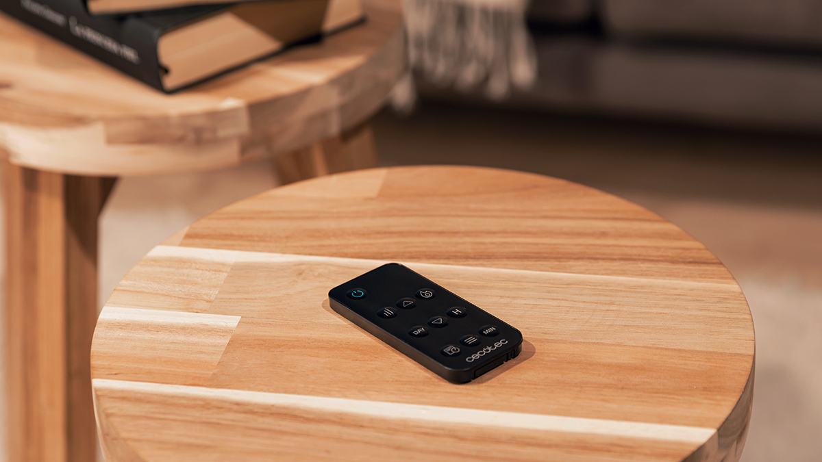 Un mando a distancia negro está sobre una mesa de madera. Otra mesa con libros en el fondo.