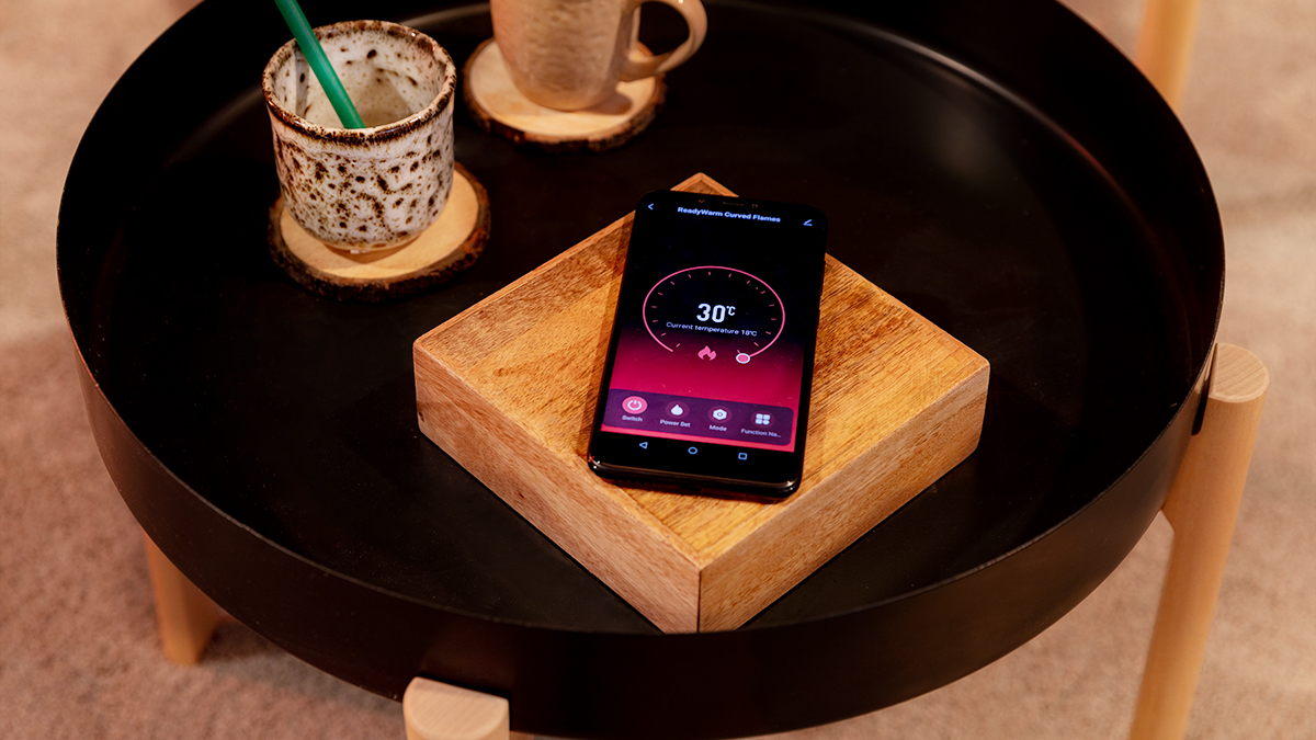 Un teléfono que muestra 30 grados está sobre un bloque de madera, en una mesa negra con una taza, un posavasos y una taza.