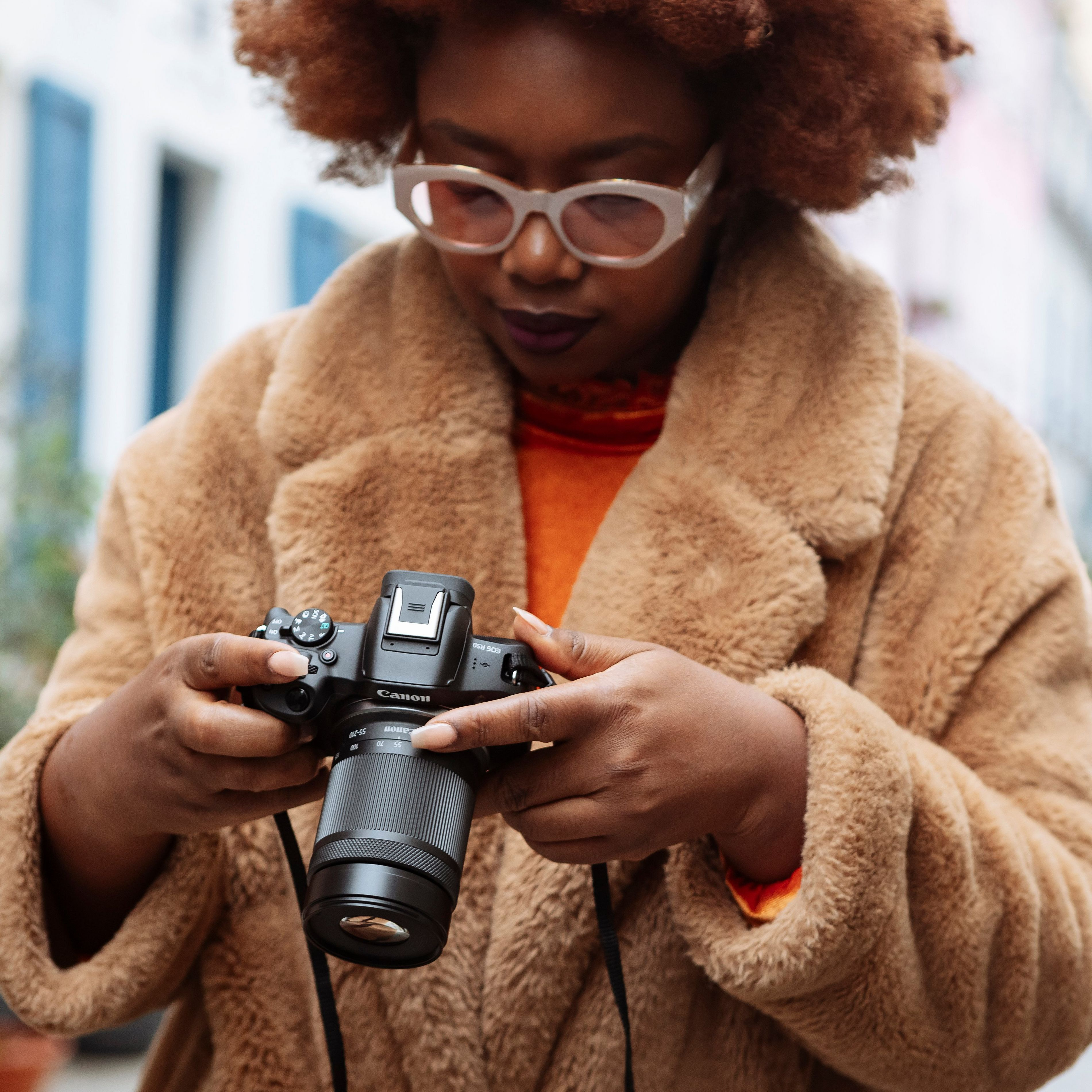 Une femme tenant un appareil photo. Elle a des lunettes et un manteau marron. L'appareil photo a un objectif noir.
