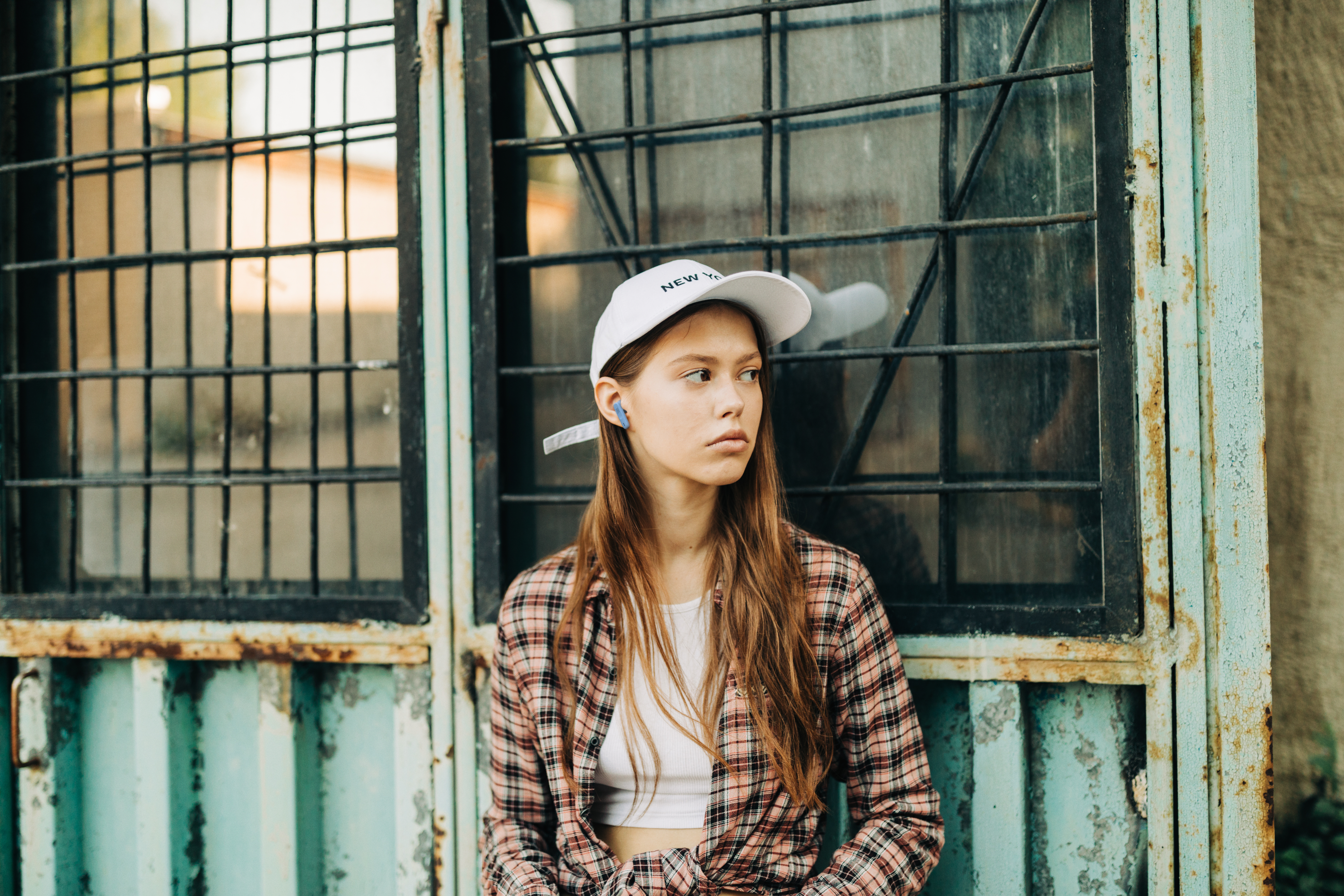 Een jonge vrouw met een witte pet en geruit overhemd staat bij een metalen poort en kijkt weg.