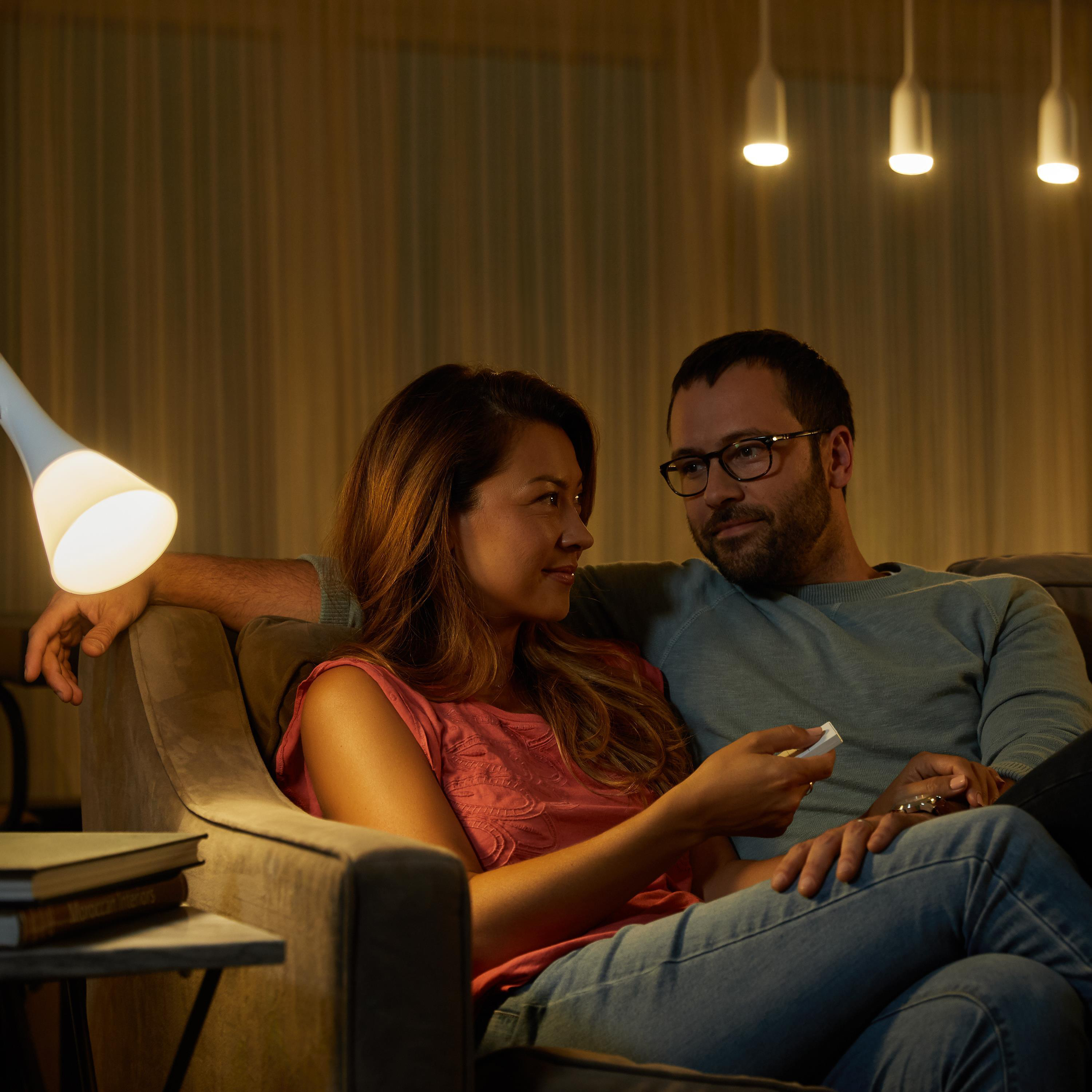 Paar auf einem Sofa, Frau schaut Mann an, der eine Fernbedienung hält. Lampe an, gemütlicher Raum.