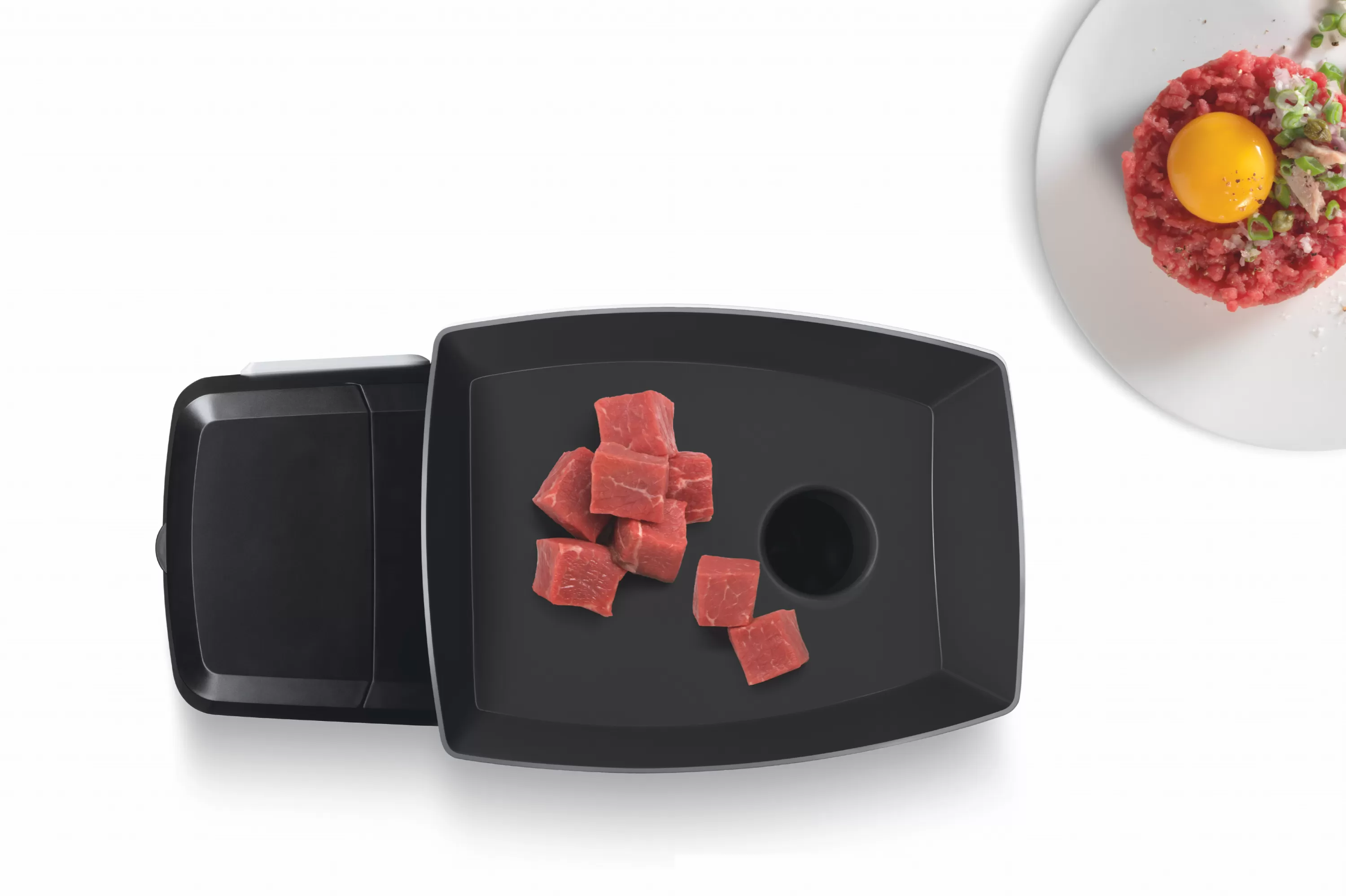 Felülnézet egy darálóról nyers húskockákkal és egy tányér elkészített steak tartárral.