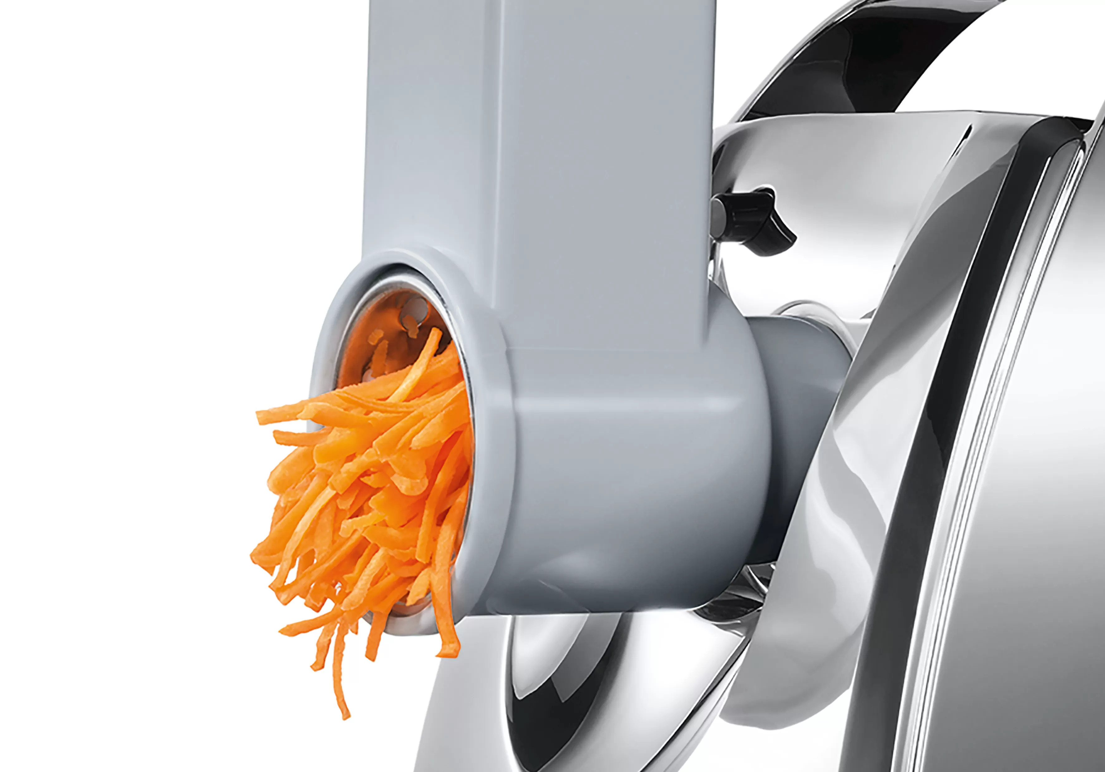 Közelkép egy élelmiszer-feldolgozóról, amely sárgarépát reszel. Narancssárga sárgarépa-forgácsok jönnek ki.