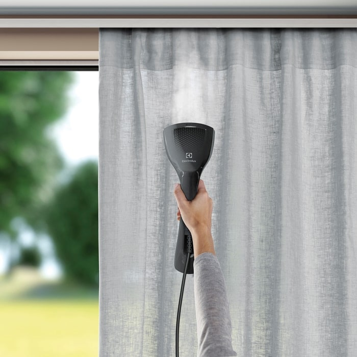 Egy kéz szürke függönyt gőzöl egy fekete Electrolux gőzölővel. Gőz emelkedik. Ablak és zöld háttérben.