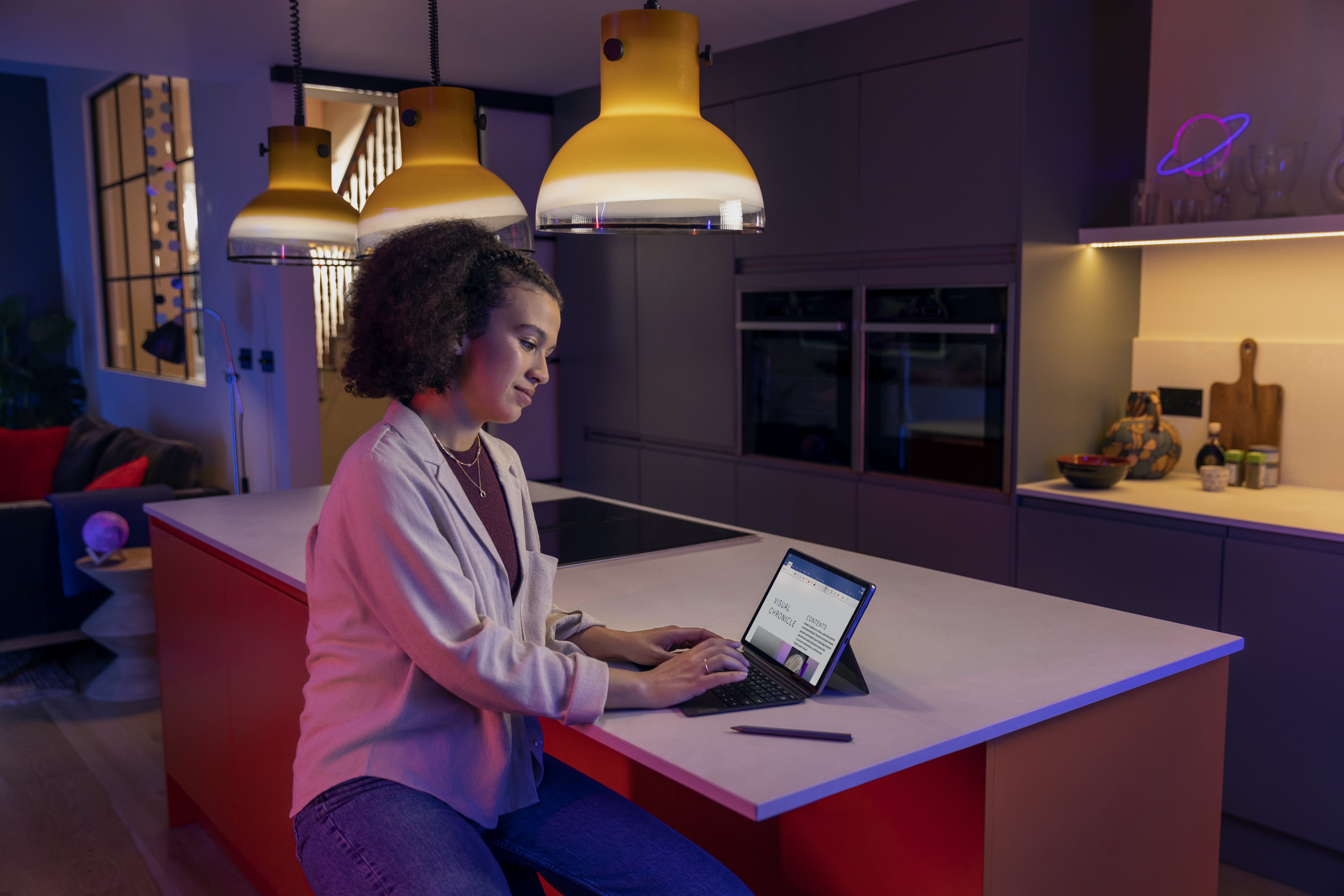 Eine Frau arbeitet an einem Laptop auf einer Kücheninsel. Drei Pendelleuchten hängen darüber. Lila Neonlicht.