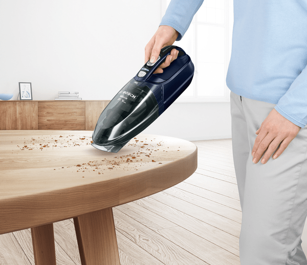 Bir kişinin eli, Bosch Move 20V el tipi elektrikli süpürgesini kullanıyor. Bir miktar döküntü ile ahşap bir masanın üzerinde.