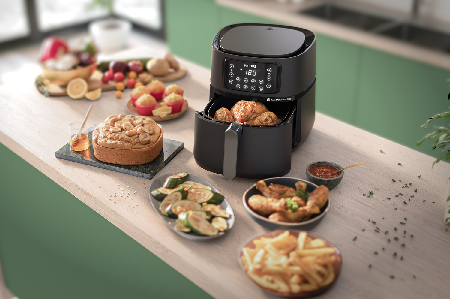 Een airfryer met eten eromheen, waaronder kip, friet en gebak. Groene achtergrond.