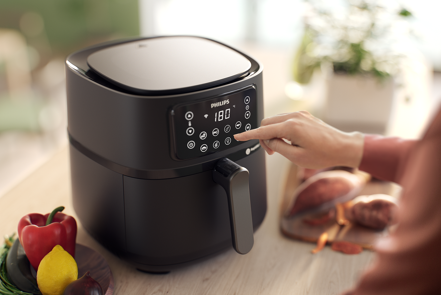 Een zwarte Philips airfryer. Een hand raakt het bedieningspaneel aan. Groenten en fruit liggen in de buurt.