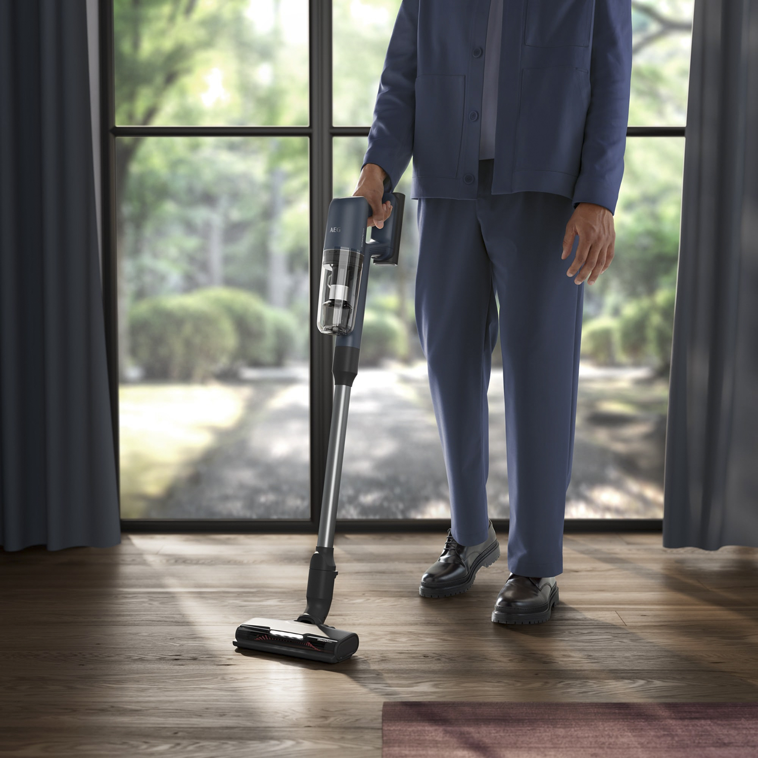 Persoon in blauw houdt een draadloze stofzuiger vast, in een kamer met een raam en bomen buiten.