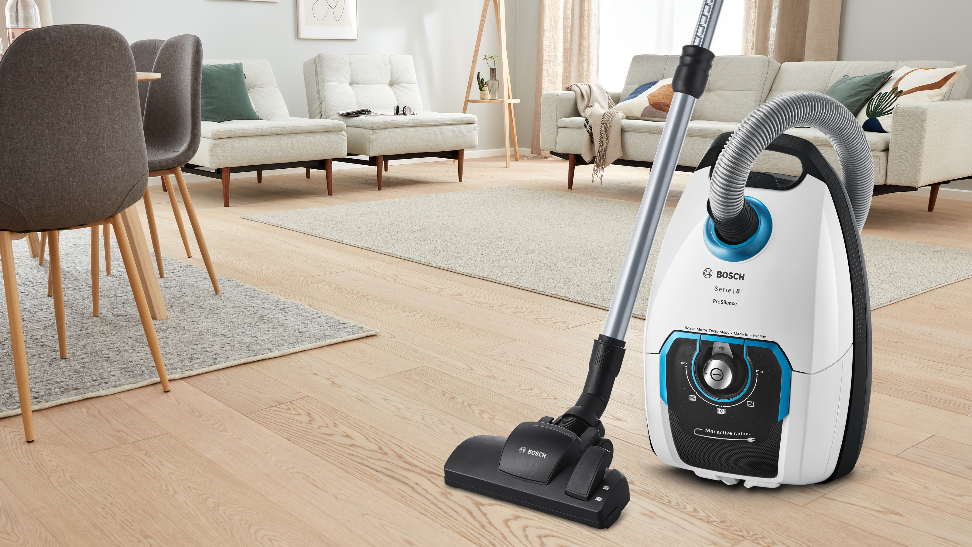 Ein Bosch-Staubsauger mit langem Schlauch steht auf Parkett in einem Wohnzimmer.