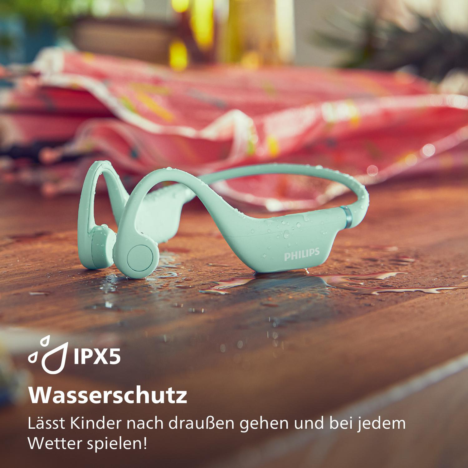 Mintgrüne Philips-Kopfhörer auf nasser Oberfläche. Text: Wasserdicht. Für Kinder draußen spielen.