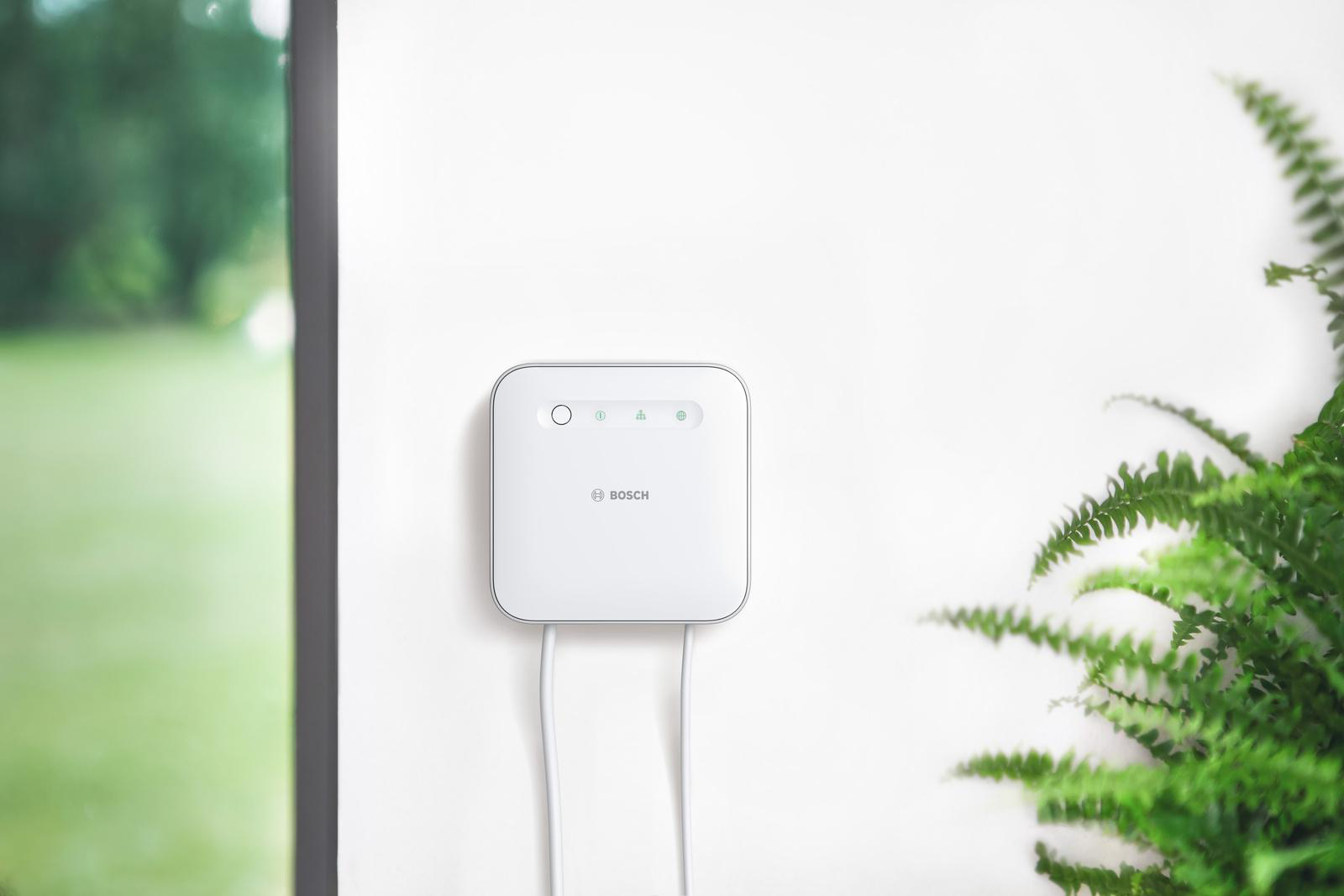 Ein weißes Bosch Smart Home-Gerät ist an einer weißen Wand montiert, mit einer grünen Pflanze im Hintergrund.