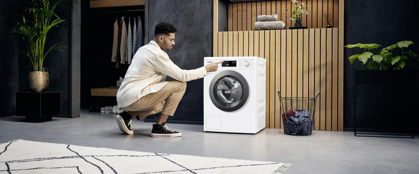Ein Mann in weißem Hemd und Hose steht neben einer weißen Waschmaschine in einem modernen, dunklen Interieur. Er berührt sie.