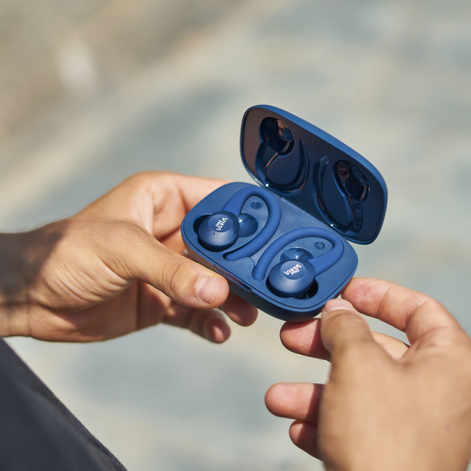 Hands holding open blue earbud case with earbuds inside. Background is blurred.