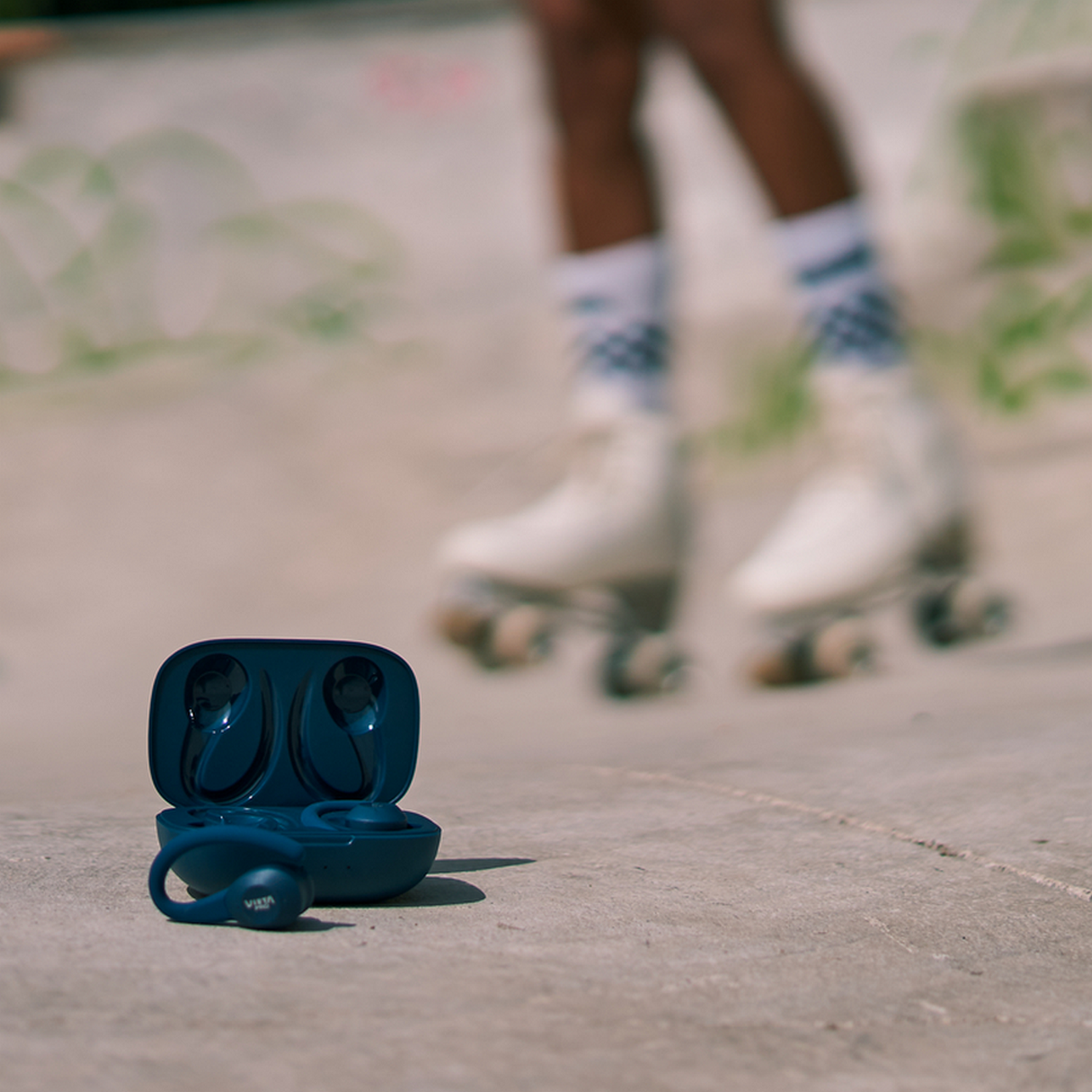 Blue earbuds and case on concrete. Blurry roller skates in background. White socks with blue patterns on feet.