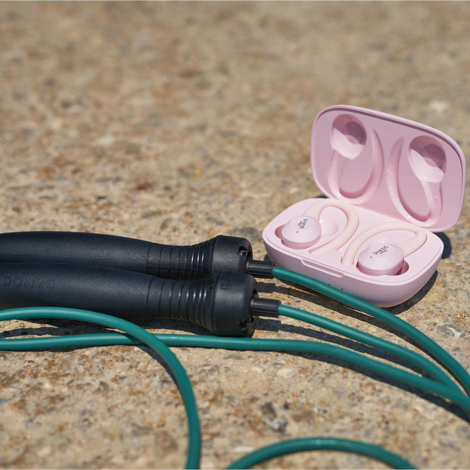 A black jump rope with green cable and pink wireless earbuds in an open case. All on a stone surface.
