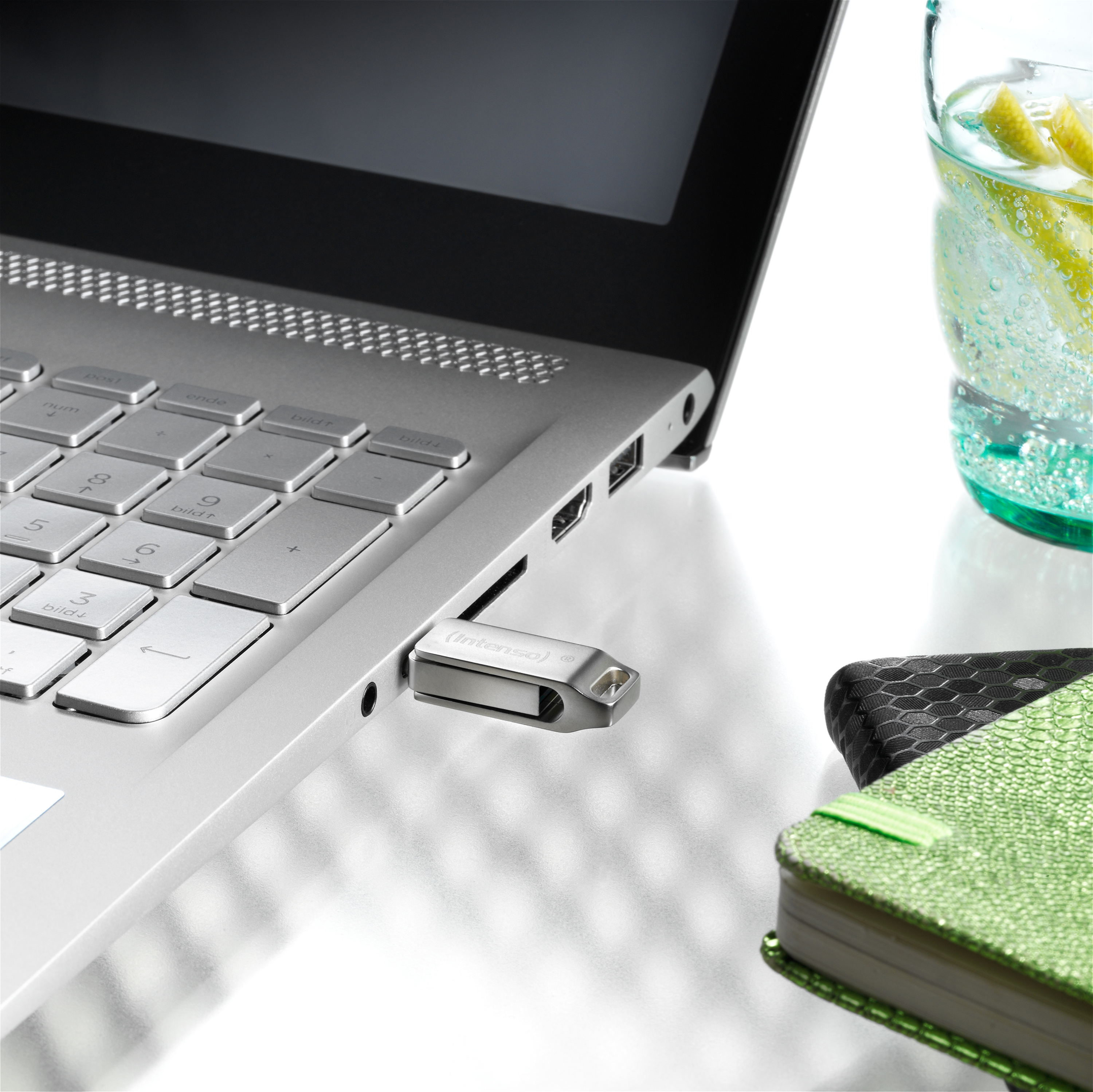 Egy laptop egy ezüst USB meghajtóval, egy citromkarikákkal teli pohár közelében.