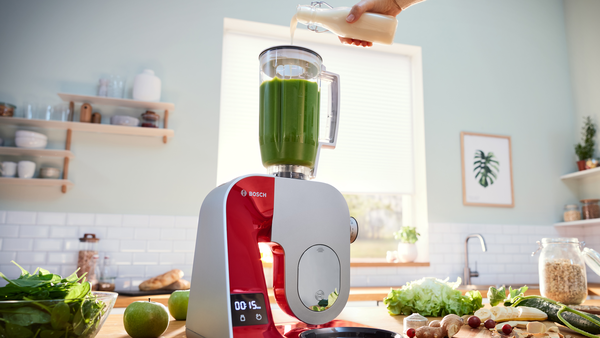 Bir blender, yeşil bir smoothie'ye sıvı döküyor. Malzemeler tezgahta.