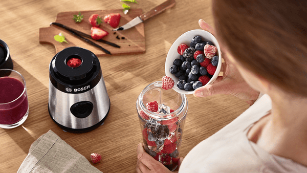 Bir kişi, ahşap bir masada smoothie ve malzemelerle birlikte, çilekleri bir blendere döküyor.