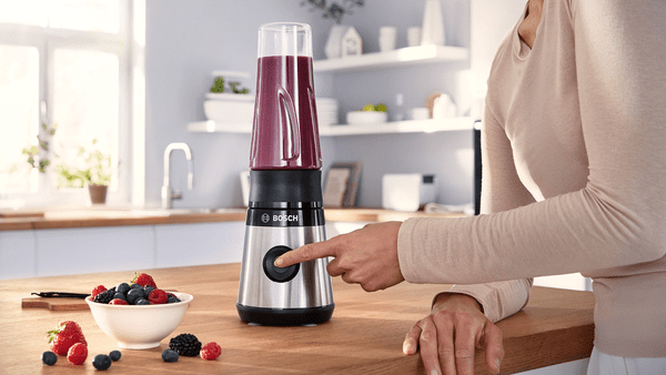 Mor bir smoothie ile gümüş ve siyah bir blender üzerinde bir düğmeye basan kadın.