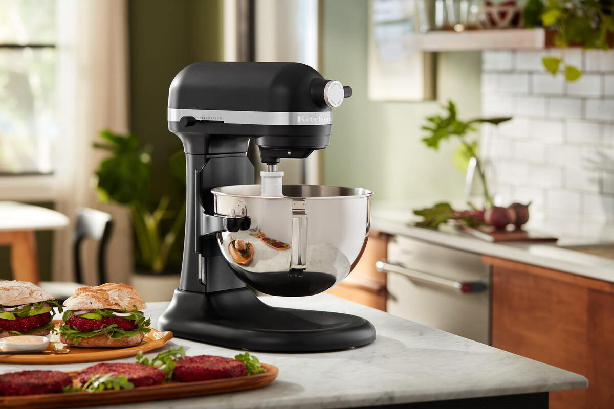 Schwarzer Küchenmixer mit Edelstahl-Schüssel, Burger und Küchenhintergrund.