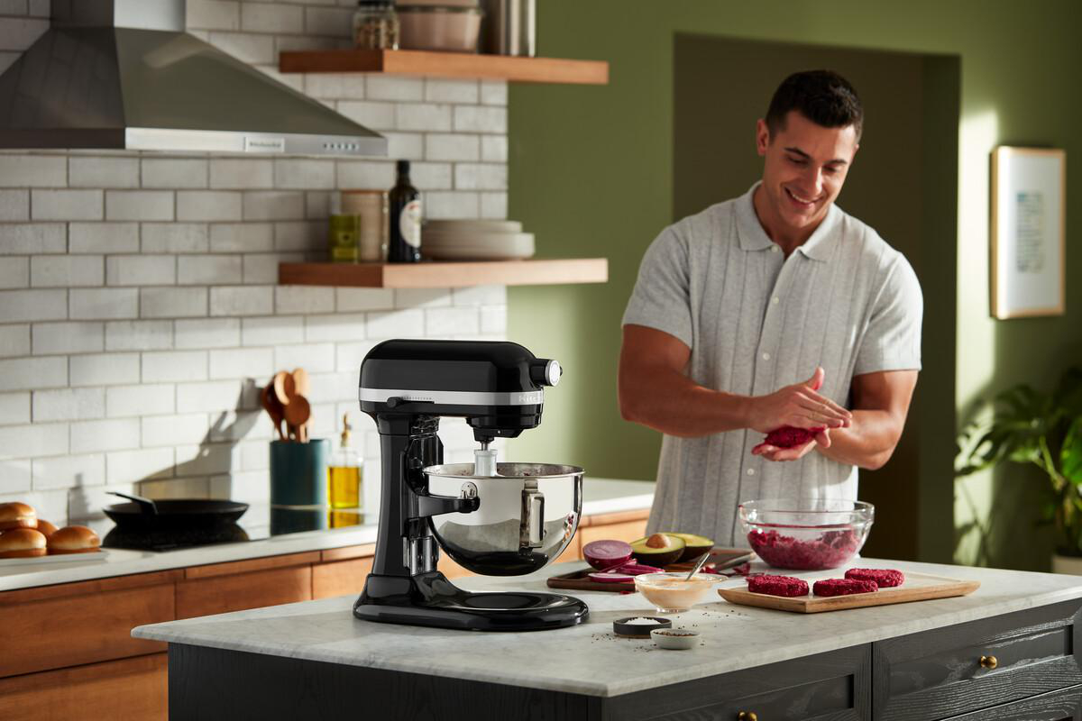 Ein Mann in einer Küche bereitet Burger zu. Ein schwarzer Mixer steht auf der Theke. Zutaten sind vorhanden.
