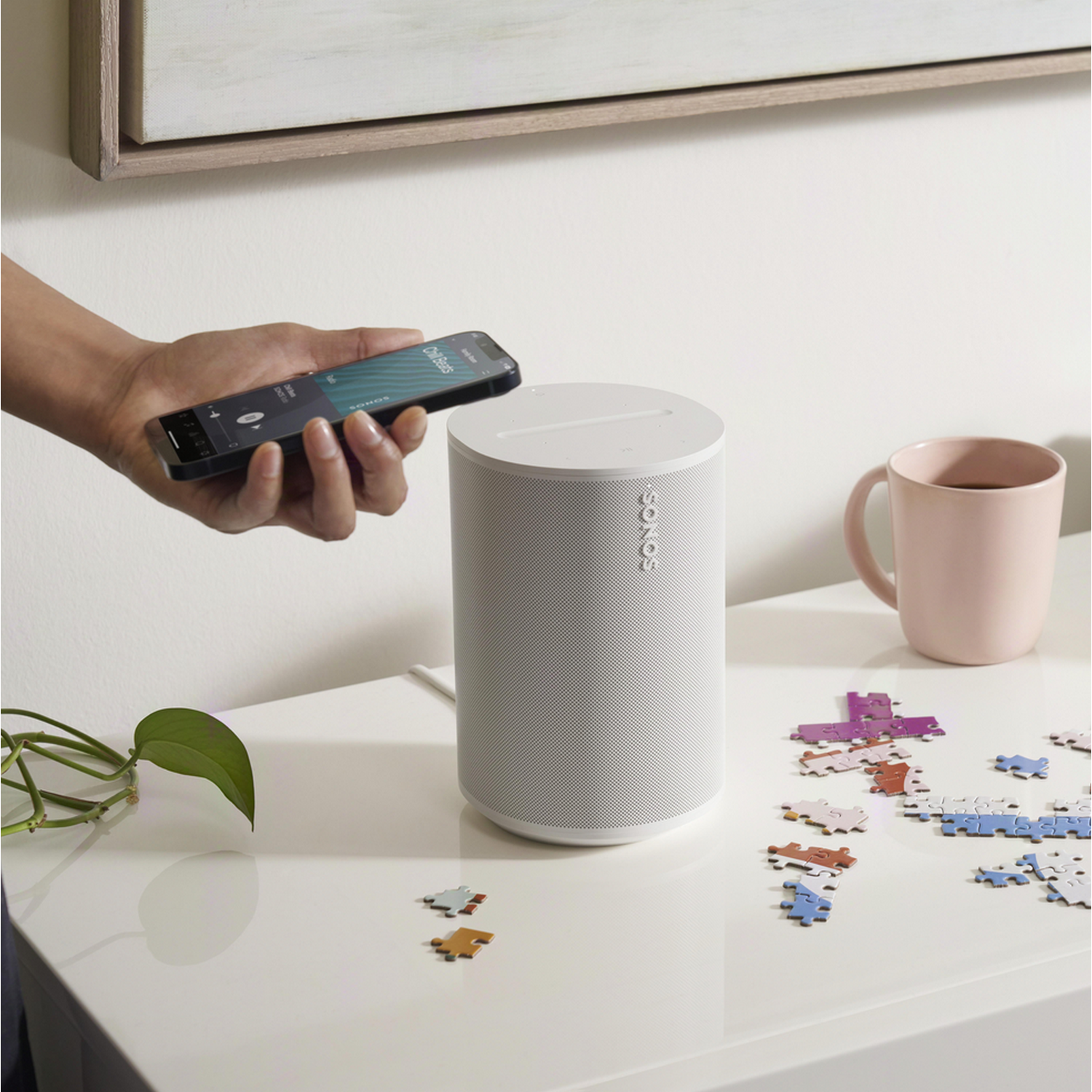 Hand holding phone near white speaker. A pink mug and a puzzle are on a white table.