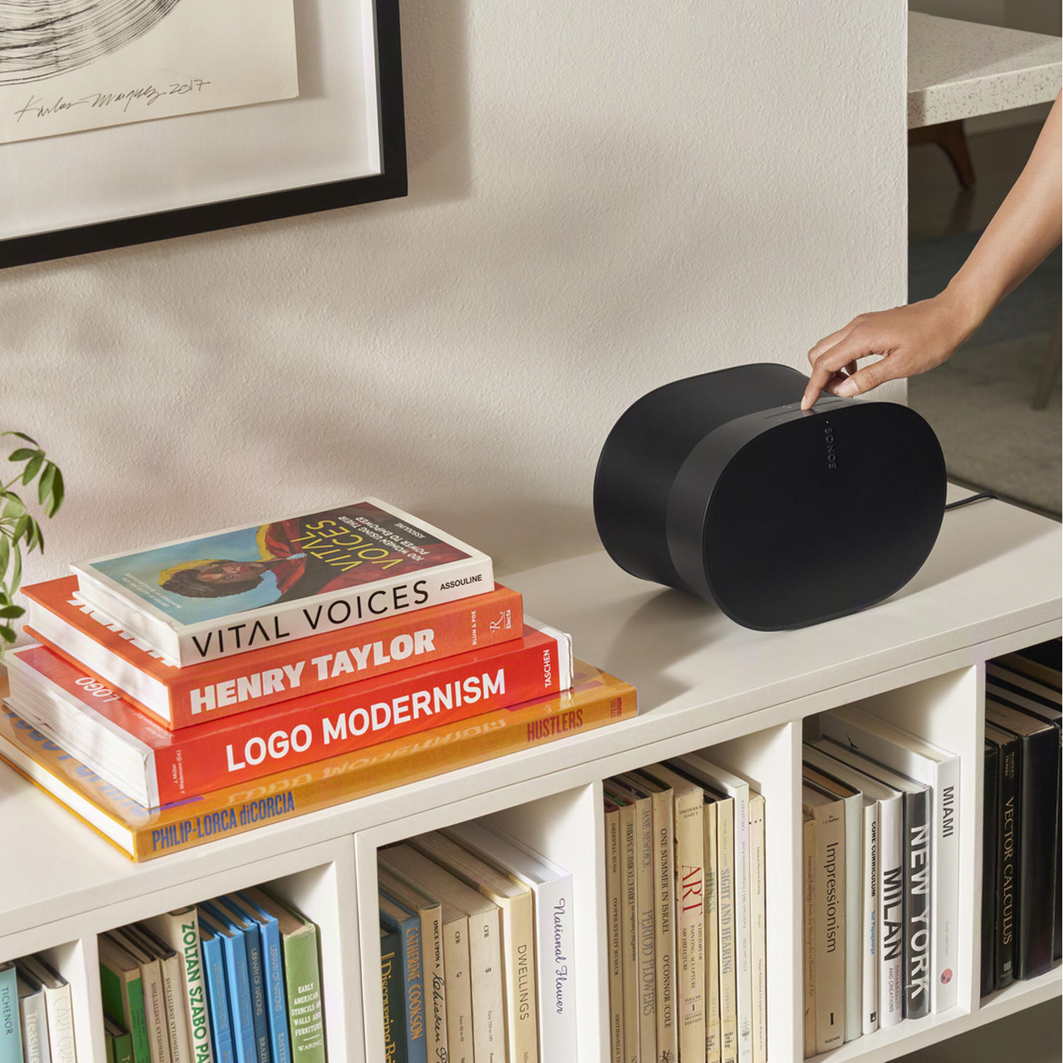 Boeken op een witte plank met een zwarte speaker. Een hand reikt naar de speaker.