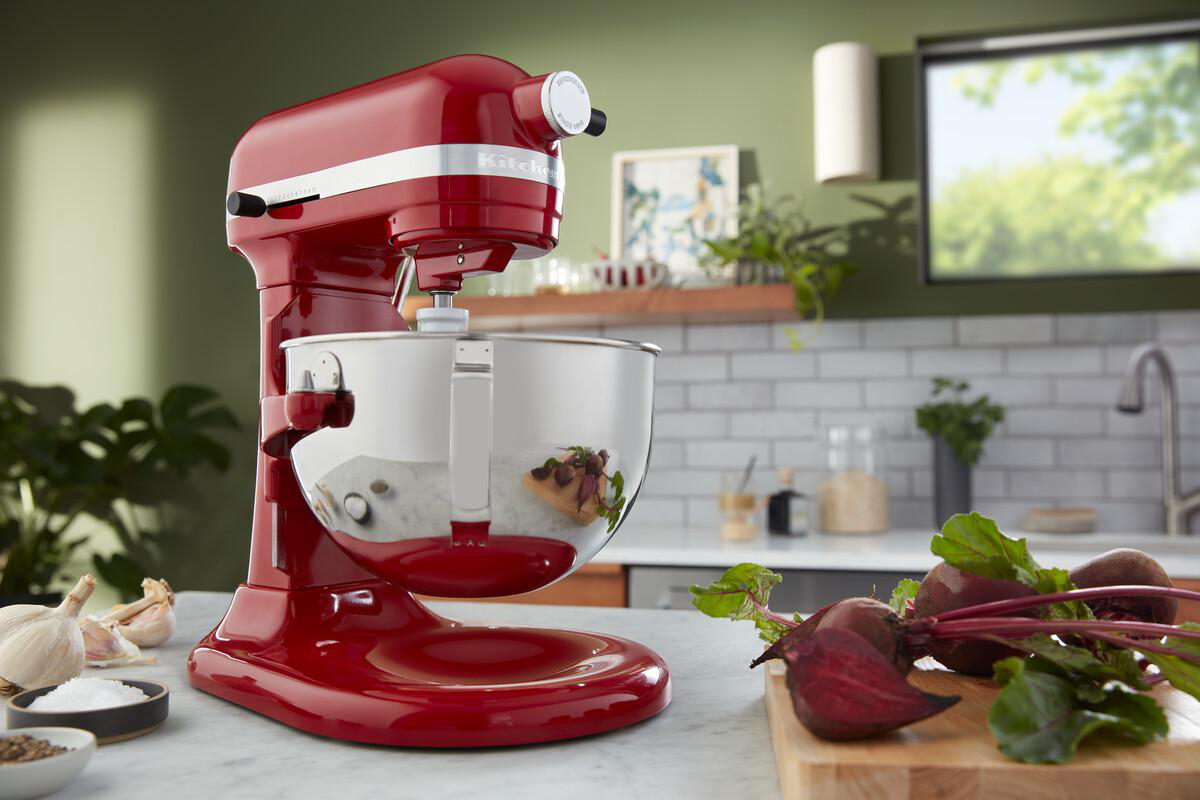 Ein roter KitchenAid-Küchenmixer mit einer silbernen Schüssel und Zutaten im Vordergrund. Die Küche ist im Hintergrund.