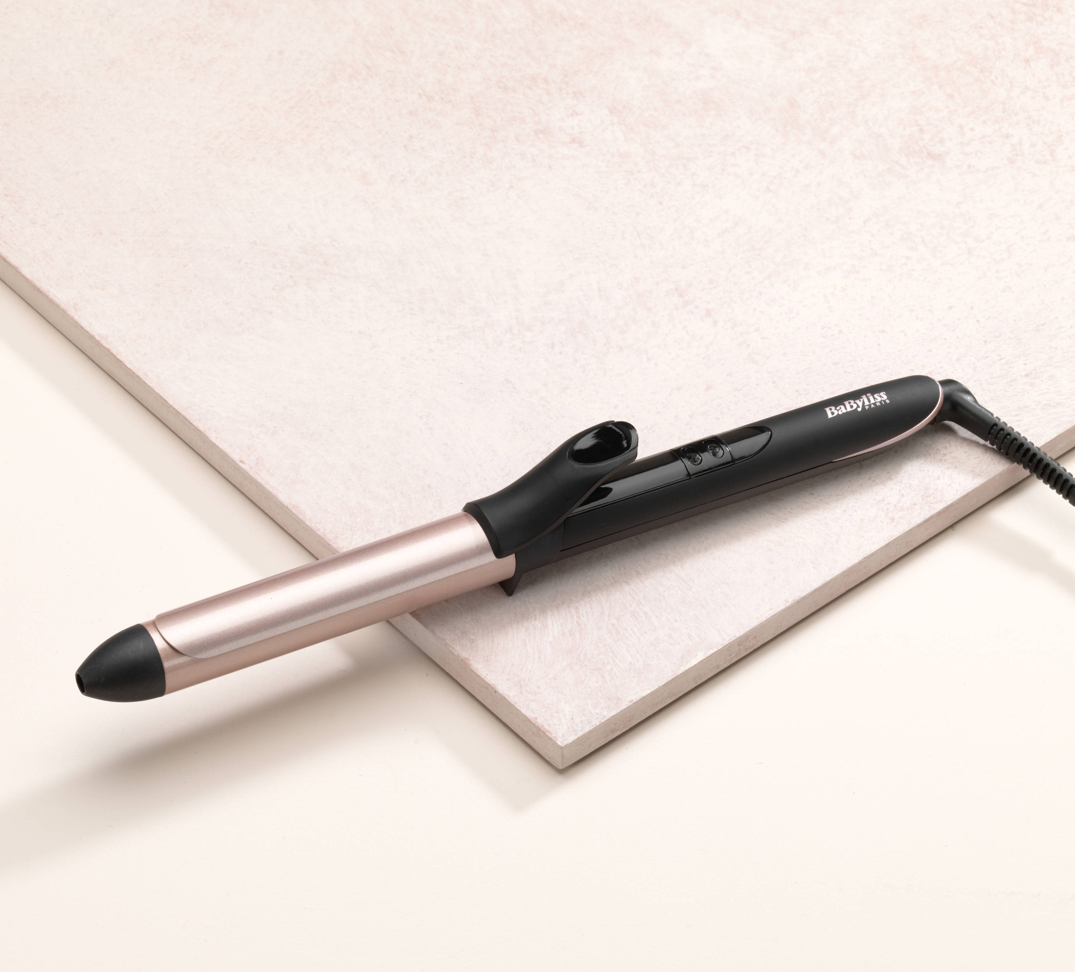 Un fer à friser noir et or rose, incliné sur une surface neutre.