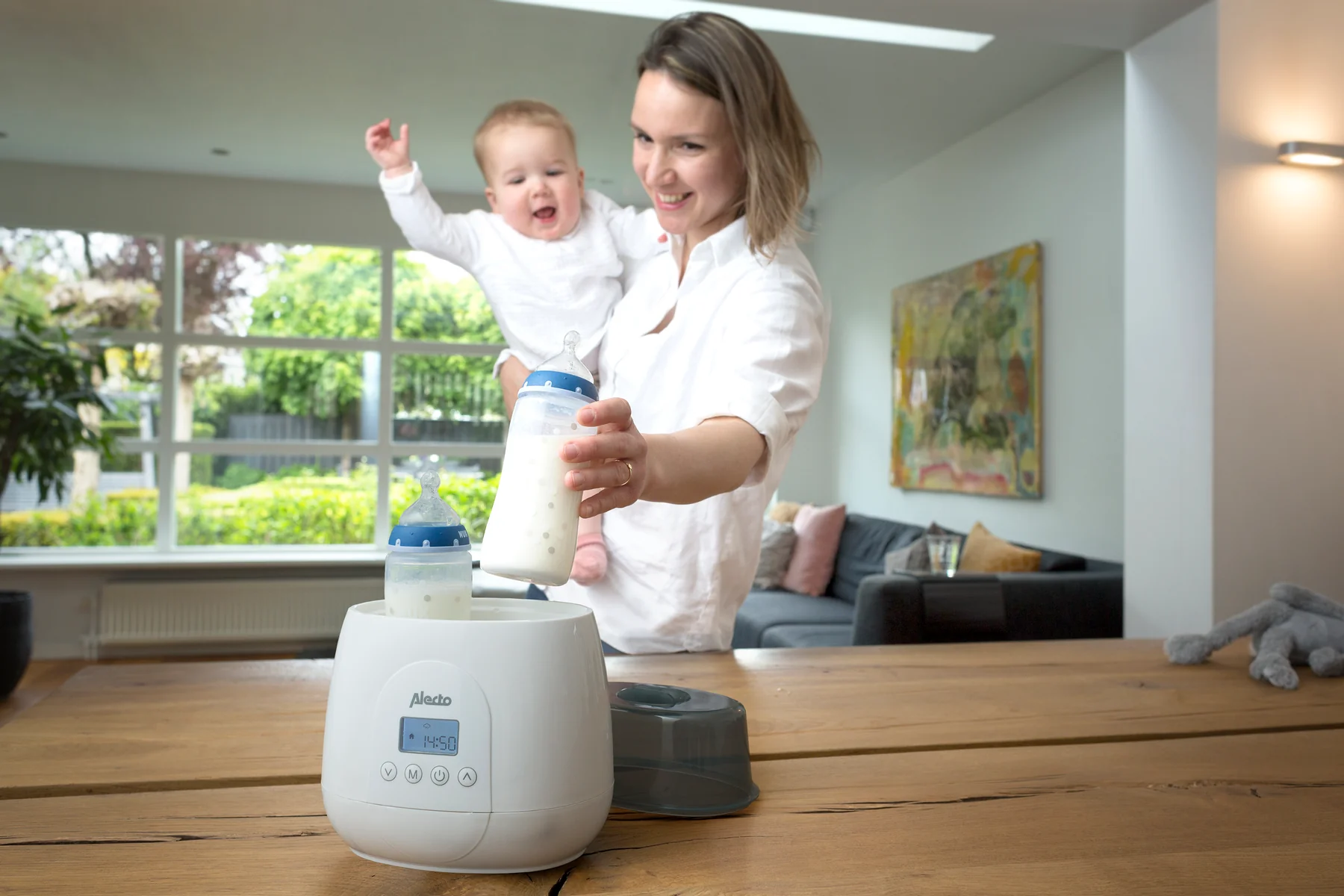Frau hält Baby, Flaschenwärmer mit Flaschen auf Holztisch, Wohnzimmer im Hintergrund.