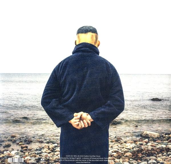 Ein Mann in einem dunkelblauen Mantel steht mit dem Rücken zur Kamera und blickt auf das Meer.