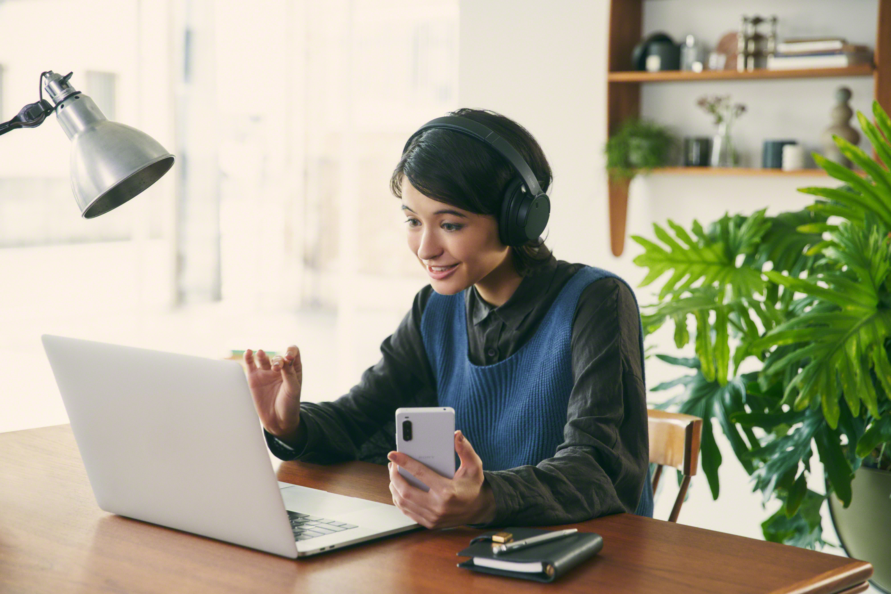 Vrouw in een videogesprek. Ze houdt een telefoon vast en kijkt naar een laptop op een houten bureau.