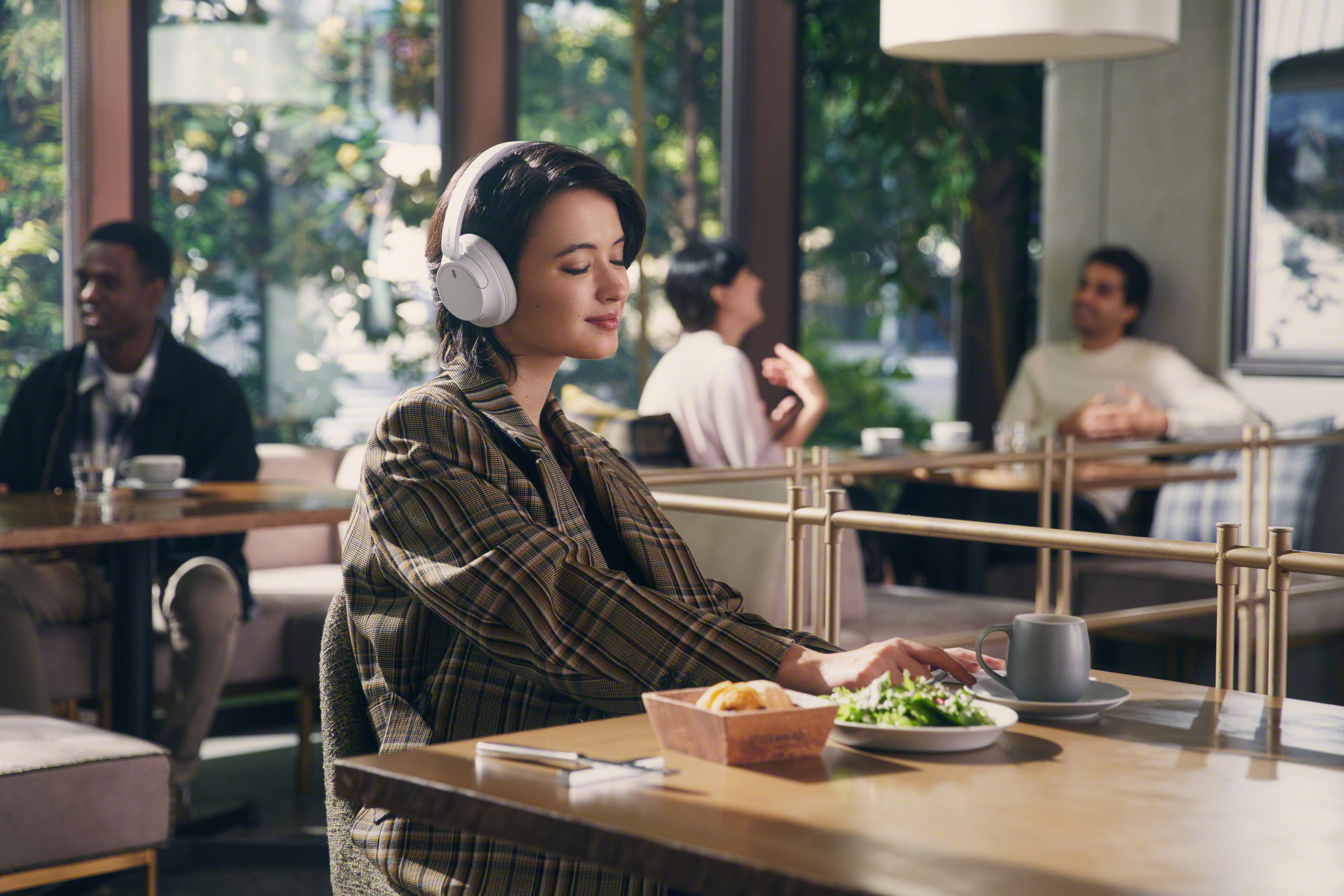 Une femme portant un casque blanc est assise à une table dans un café. Elle regarde vers le bas et travaille sur un ordinateur portable.