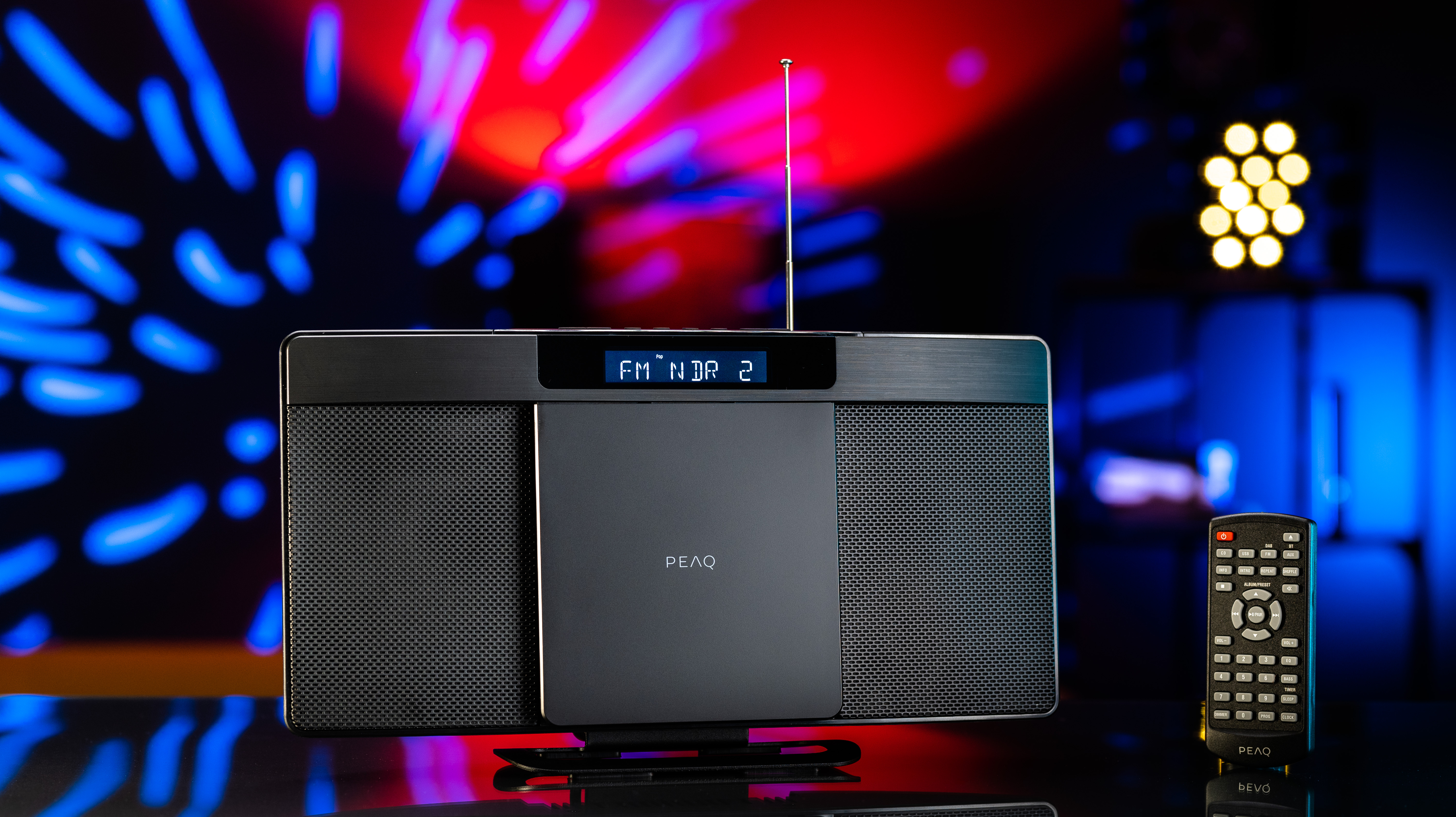 Schwarzes Radio mit Display. Fernbedienung rechts. Dunkler Hintergrund mit bunten Lichtern.
