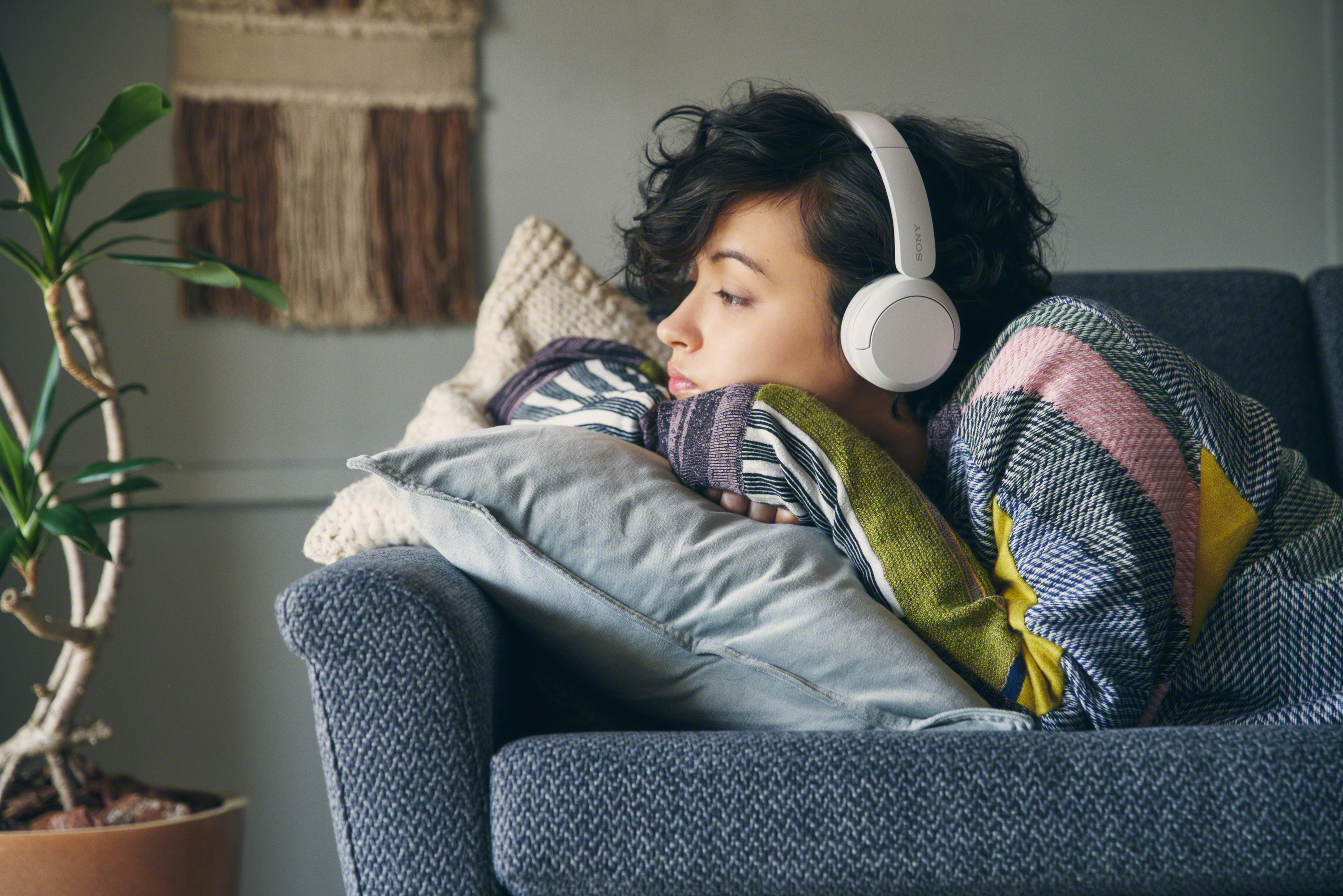 Femme sur un canapé avec un casque et une couverture, regardant pensivement vers la gauche.