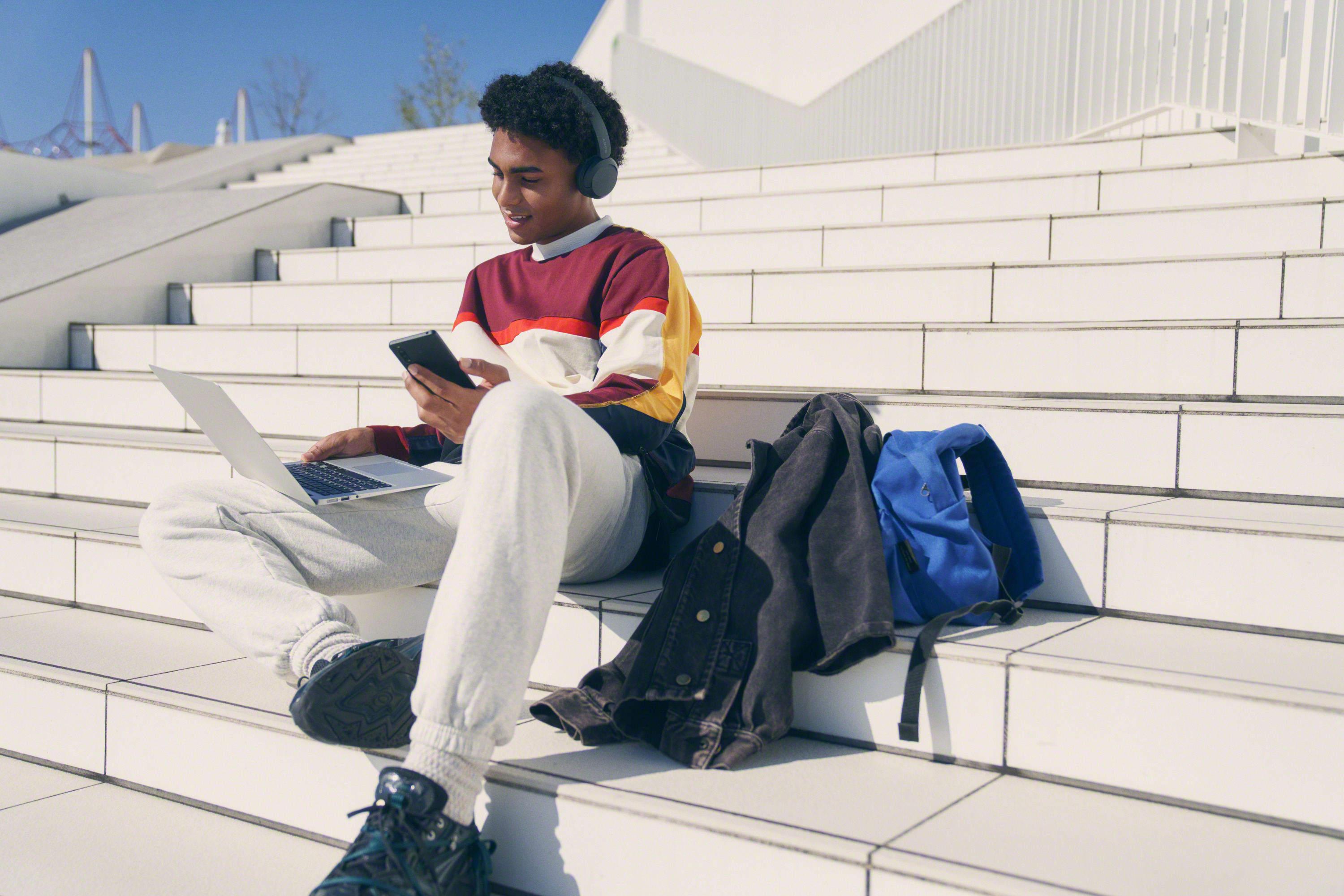 Young man sitting on stairs, using phone and laptop. Backpack, headphones, and jacket nearby.