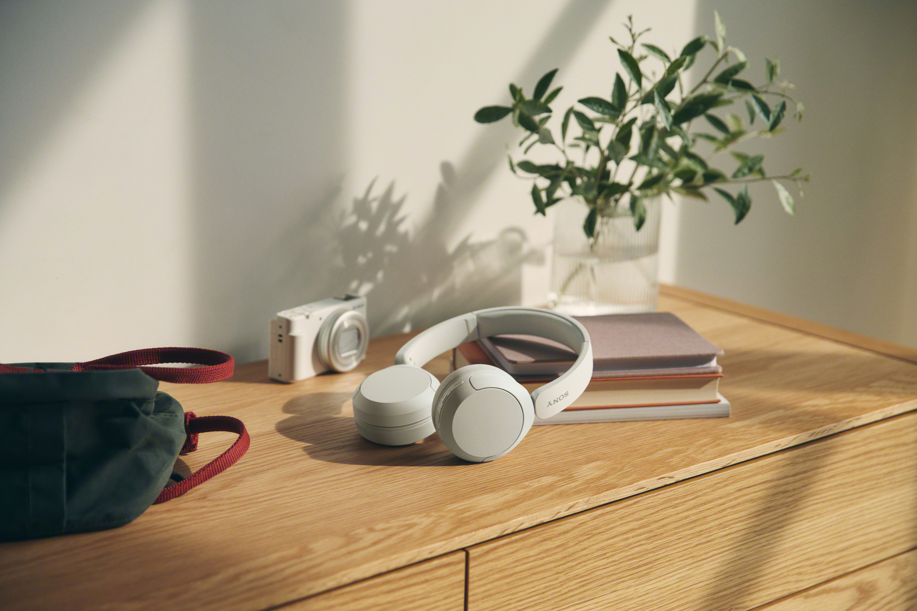 Un bureau en bois avec un casque, un appareil photo, un journal et une plante dans un vase en verre. Soleil.