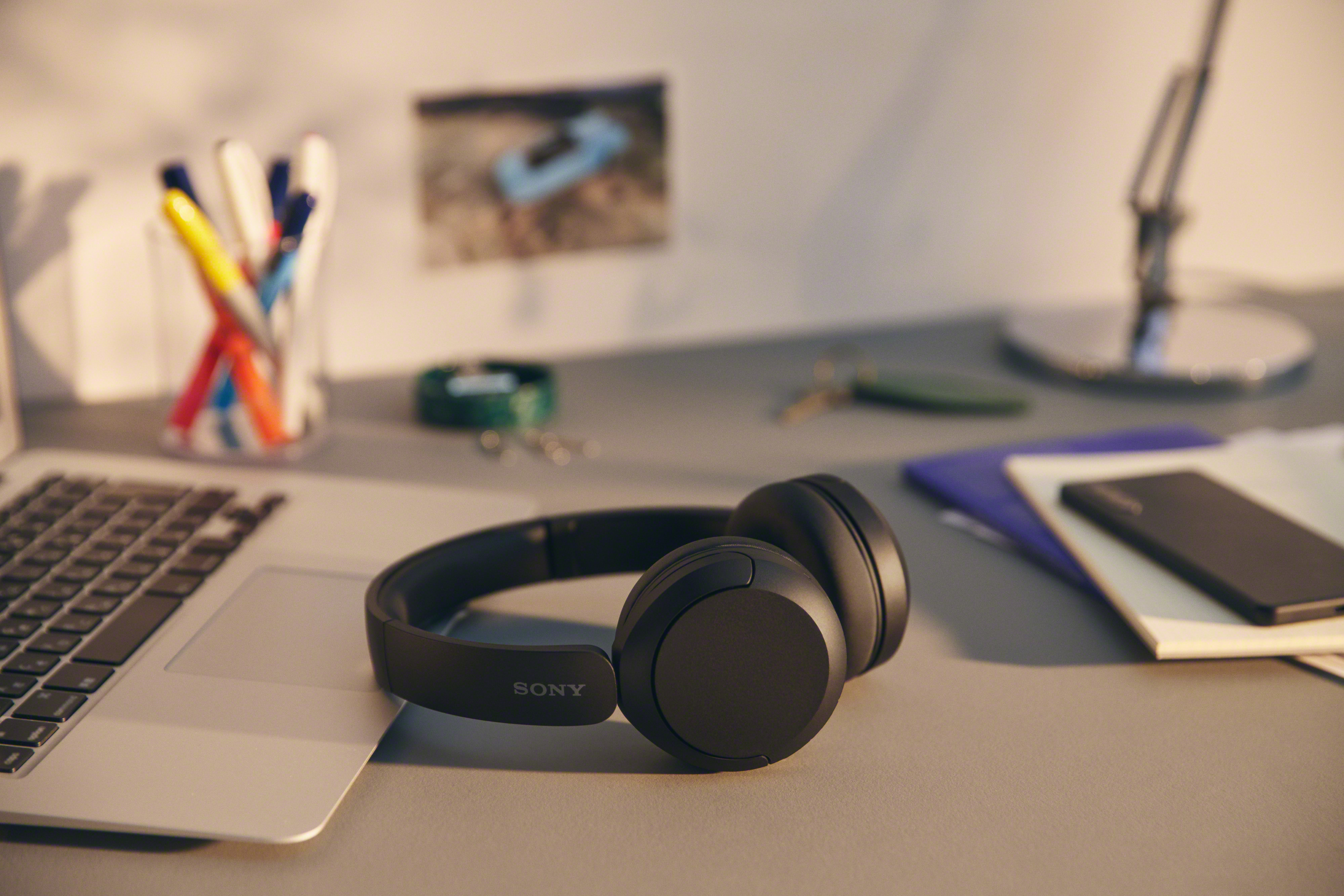 Black Sony headphones on a desk with a laptop, pens, and a picture in the background.