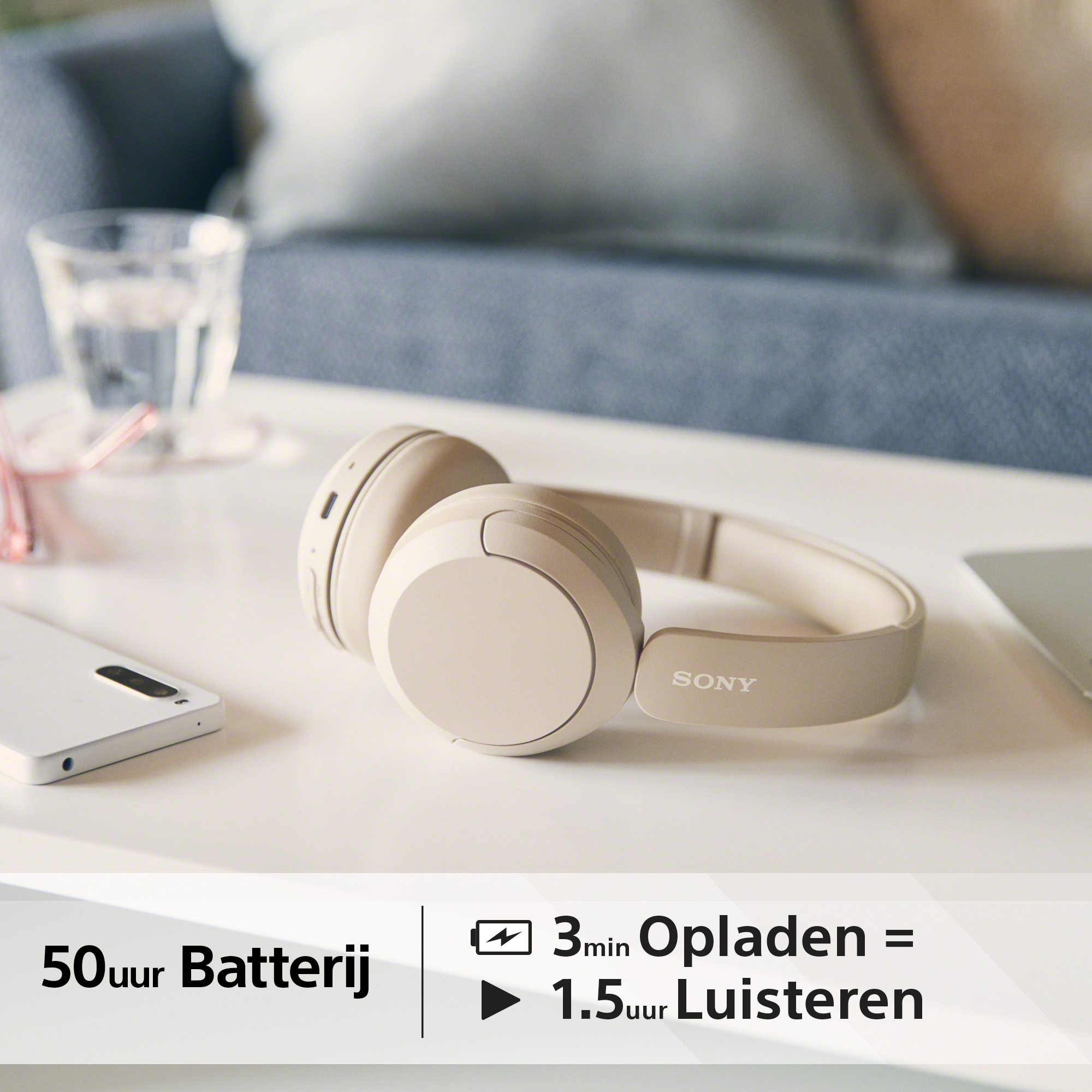 Beige koptelefoon op een tafel, naast een telefoon en een glas water. Tekst geeft de batterijduur aan.