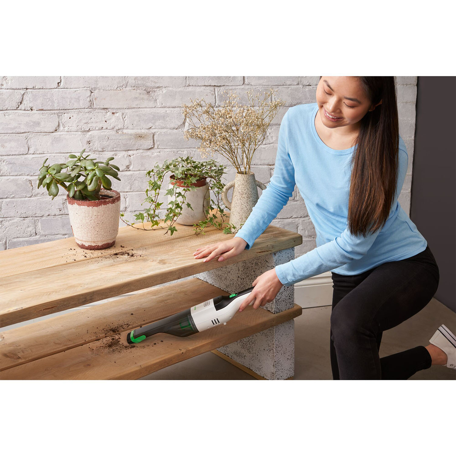 Una donna aspira un tavolo di legno con un aspirapolvere portatile. Piccole piante e fiori sullo sfondo.