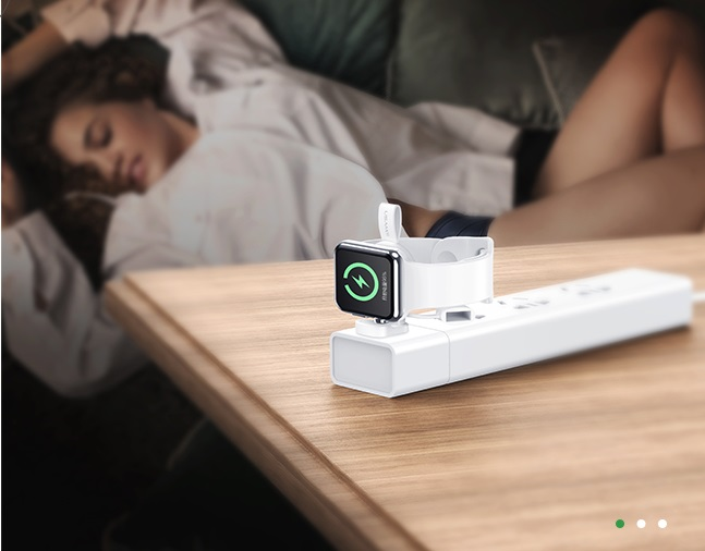 Egy Apple Watch töltődik egy fehér elosztón, a háttérben egy nő alszik a kanapén.